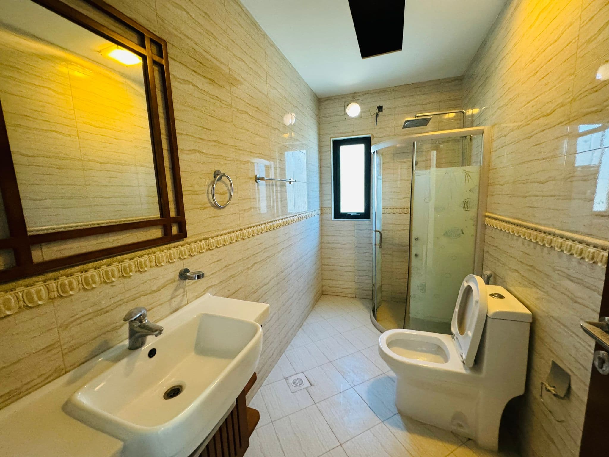 A clean bathroom with a sink, toilet, and a glass shower enclosure. Beige tiled walls and floor, a mirror above the sink, and a window for natural light.