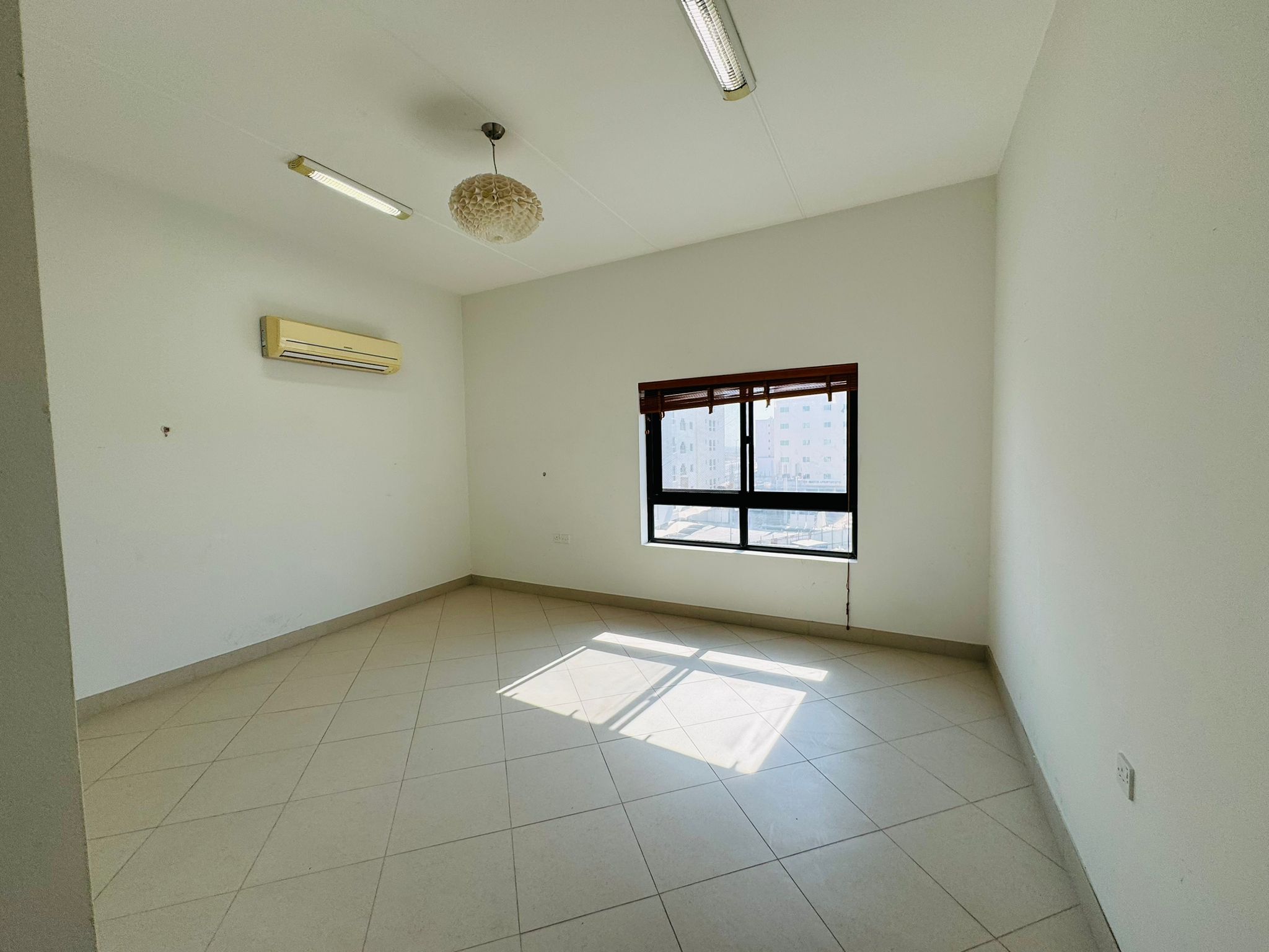 A vacant room with tiled floors, a window with blinds, an air conditioning unit, and overhead lights.