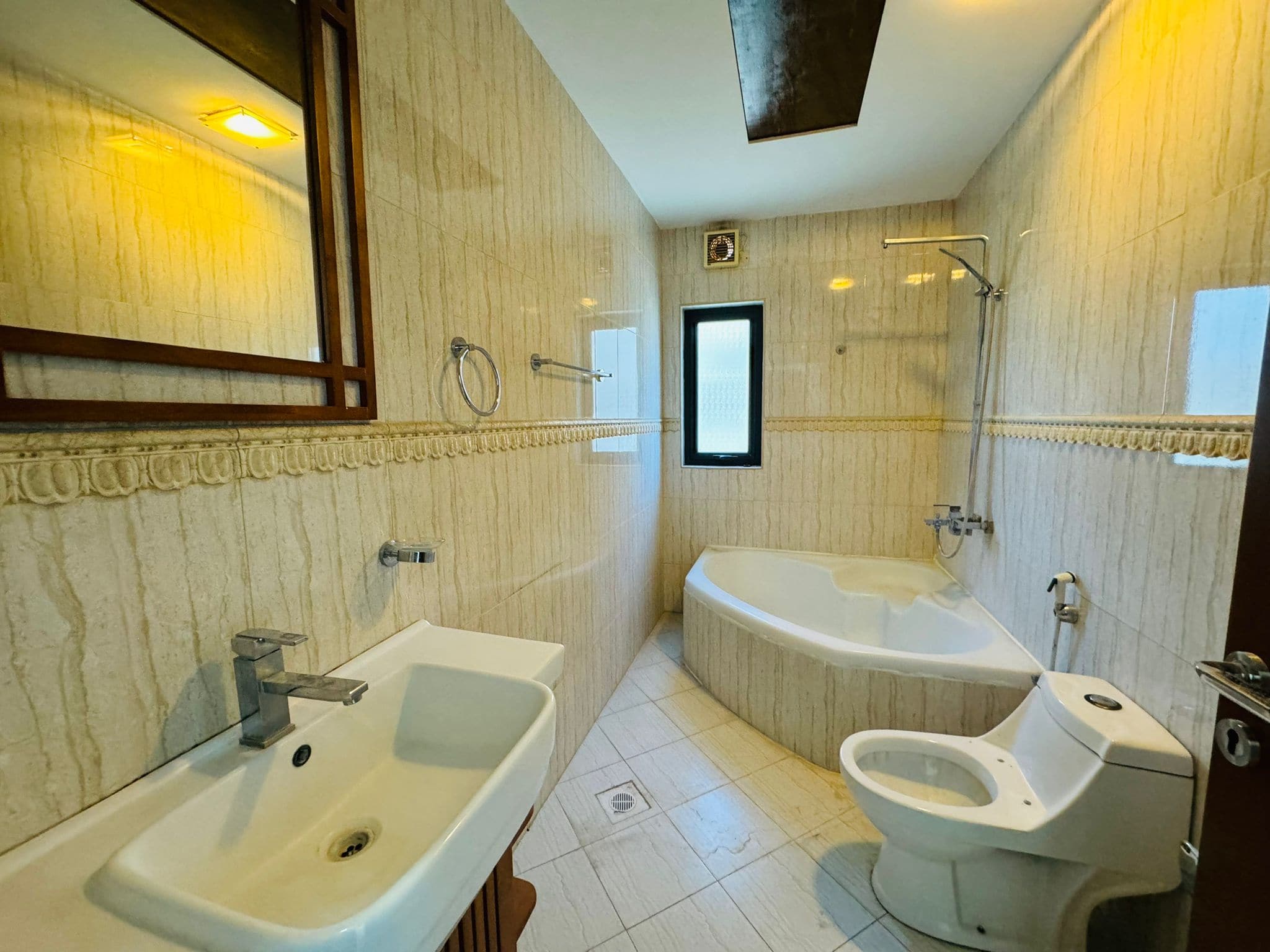 A bathroom with a sink, wall mirror, toilet, and a corner bathtub. Beige tiles cover the walls and floor. A small window is above the bathtub.