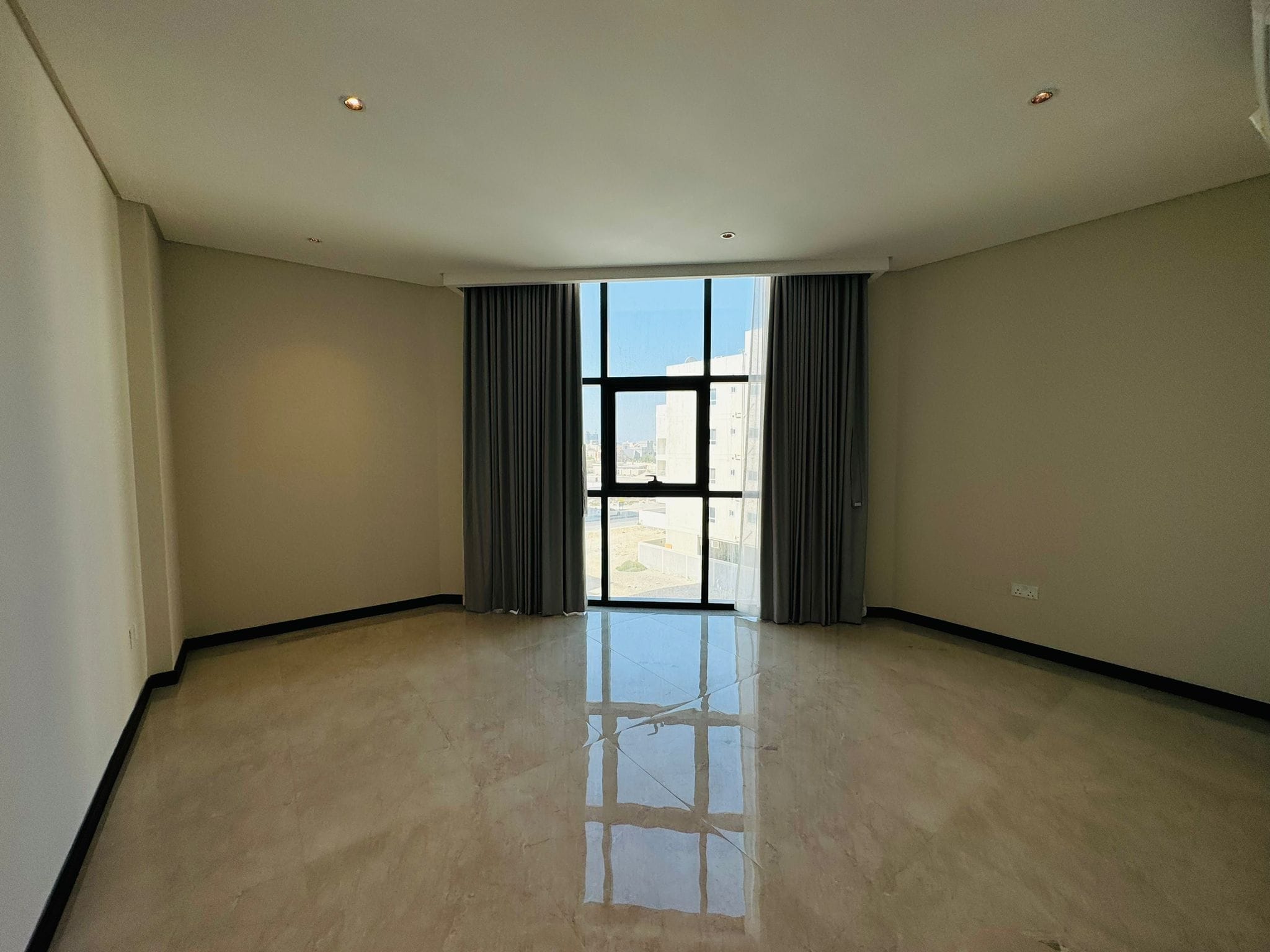 Empty room with a large window, gray curtains, and beige tiled floor. Sunlight reflects on the floor. Walls are painted light beige.