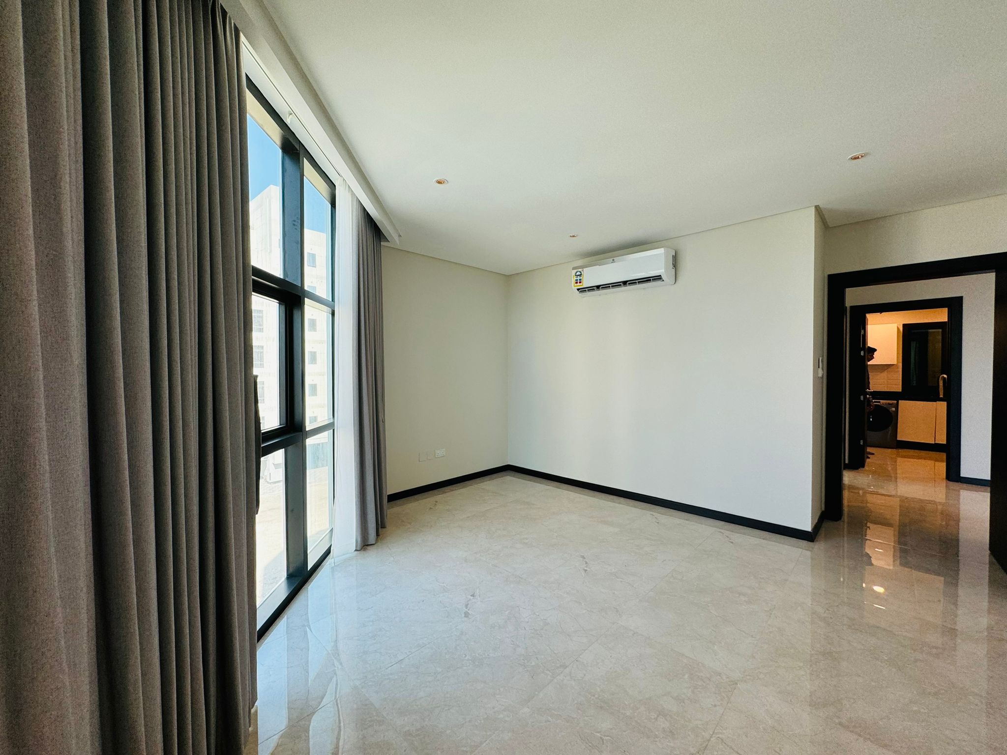 A bright, empty room with large windows, light-colored marble floors, gray curtains, and a wall-mounted air conditioning unit. A doorway leads to another area.