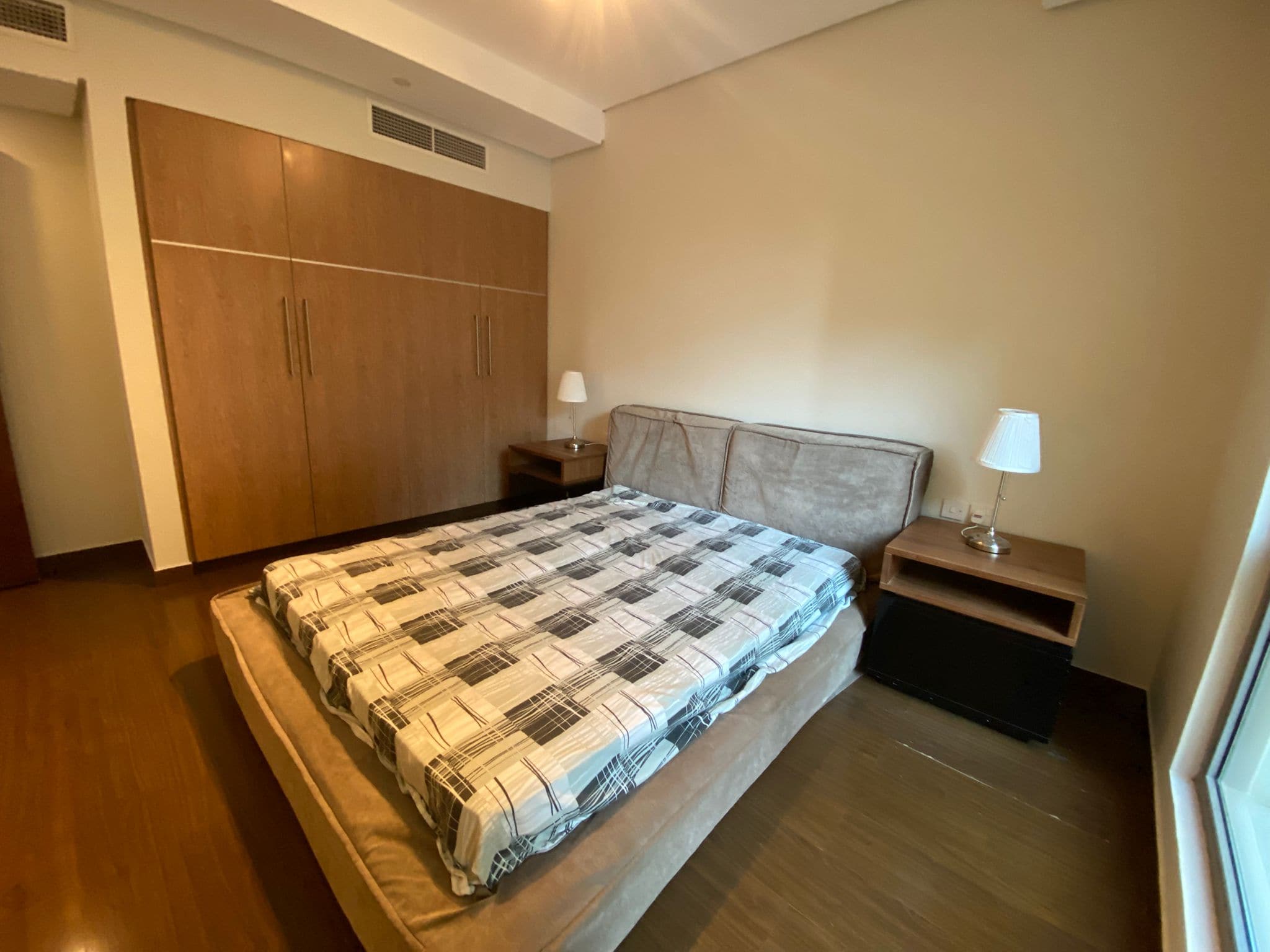 Bedroom with a double bed featuring a checkered blanket, two bedside tables with lamps, wooden floor, built-in wardrobe, and beige walls.