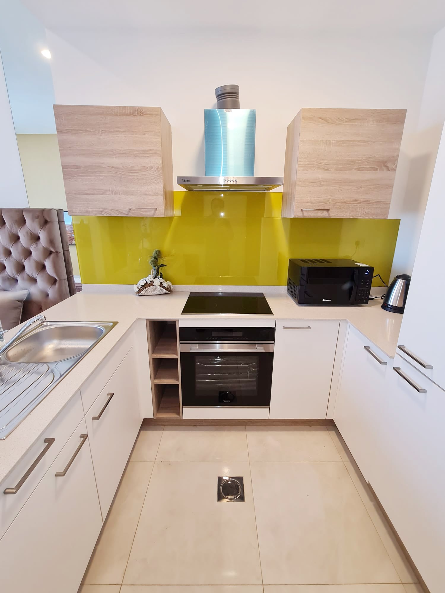 Modern kitchen with light wood cabinets, green backsplash, electric stovetop, oven, microwave, and kettle. Sink and drainer on the left. Floor has a central drain.