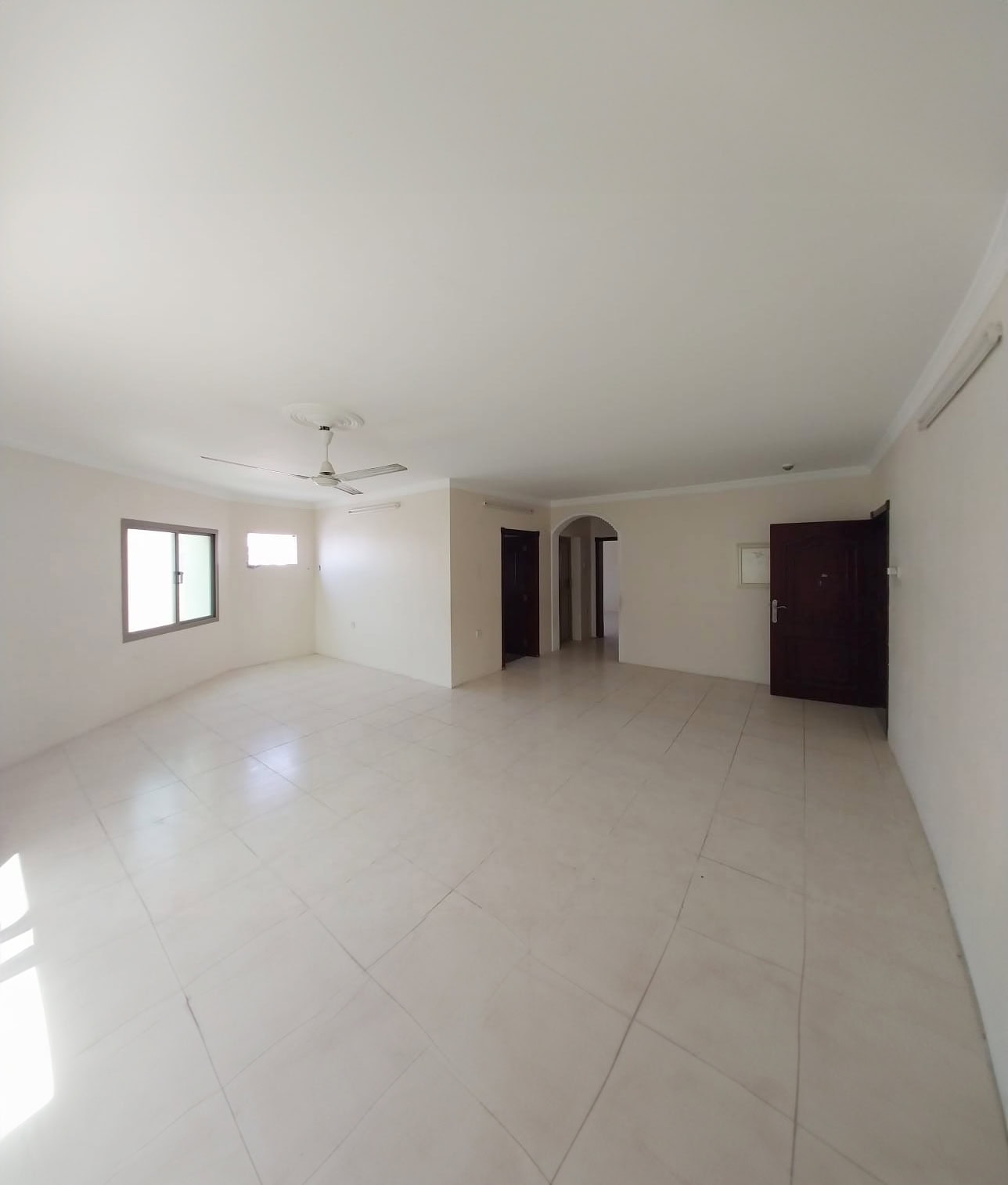 Empty room with white walls and tiled floor, featuring two doors, two windows, and a ceiling fan. Appears well-lit with natural light from the windows.