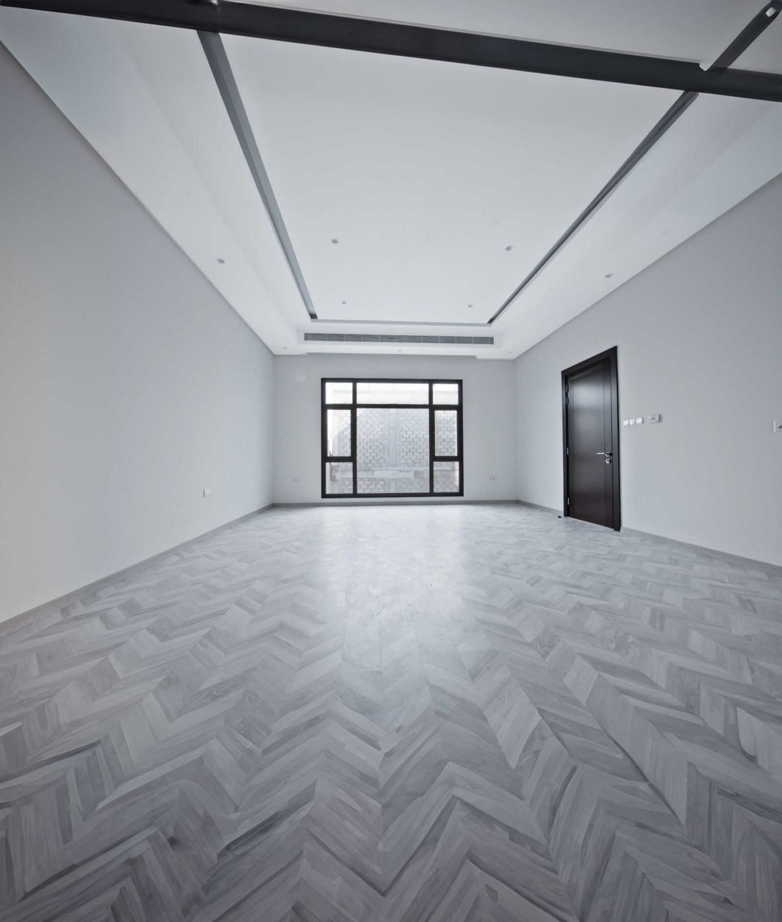 Empty room with herringbone parquet flooring, white walls, and large windows. A single dark door is on the right. Ceiling features recessed lighting and minimalist design elements.