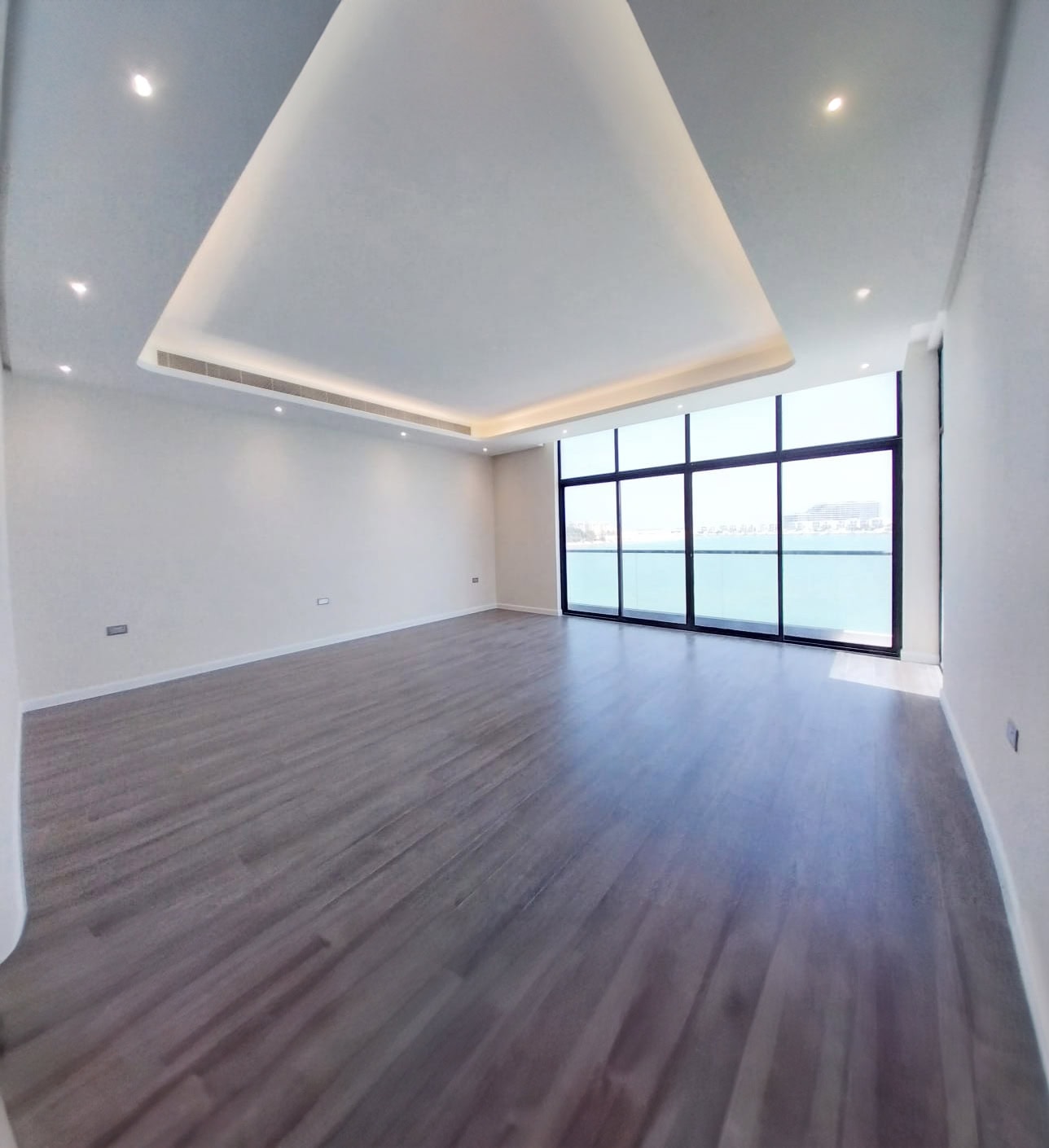 Spacious empty room with wooden flooring and large floor-to-ceiling windows revealing a water view. Ceiling features recessed lighting.