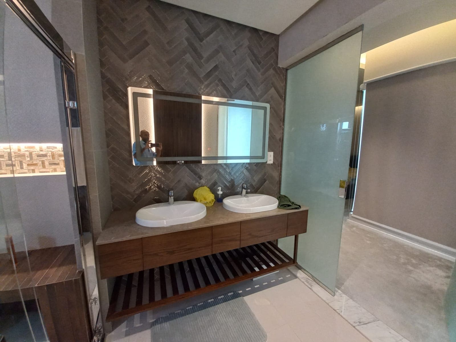 Modern bathroom with a double sink vanity, large mirror, herringbone tile backsplash, and glass shower. A man is reflected in the mirror taking a photo.