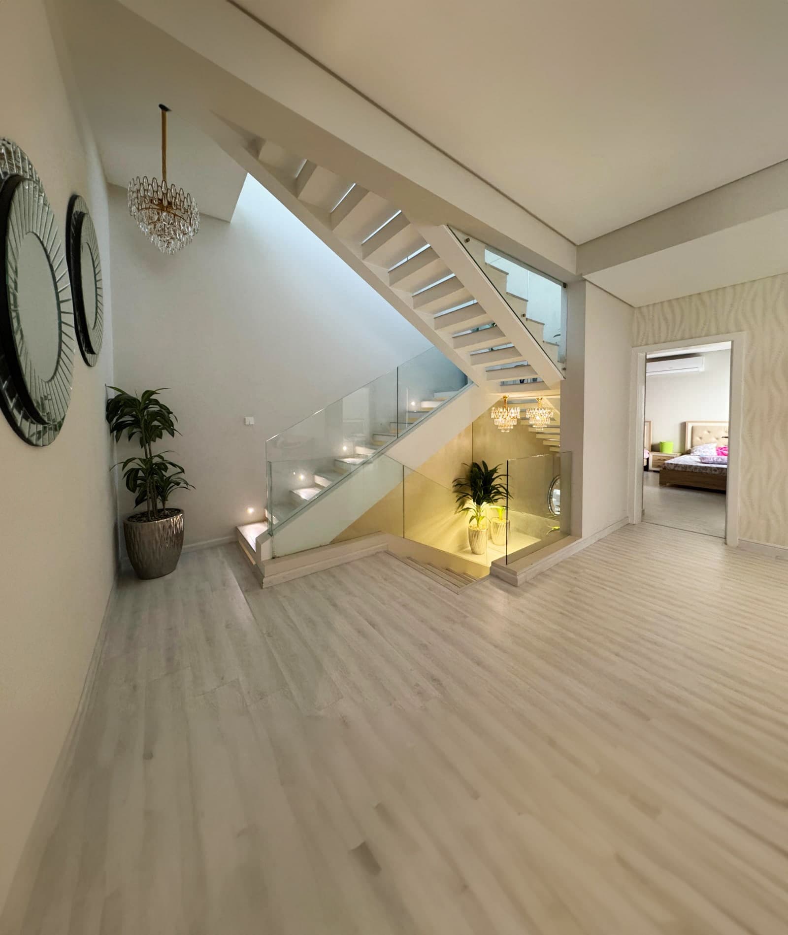 Modern hallway with light wooden flooring, a staircase with glass balustrade, potted plants, and circular wall mirrors. A bedroom is visible through an open doorway.