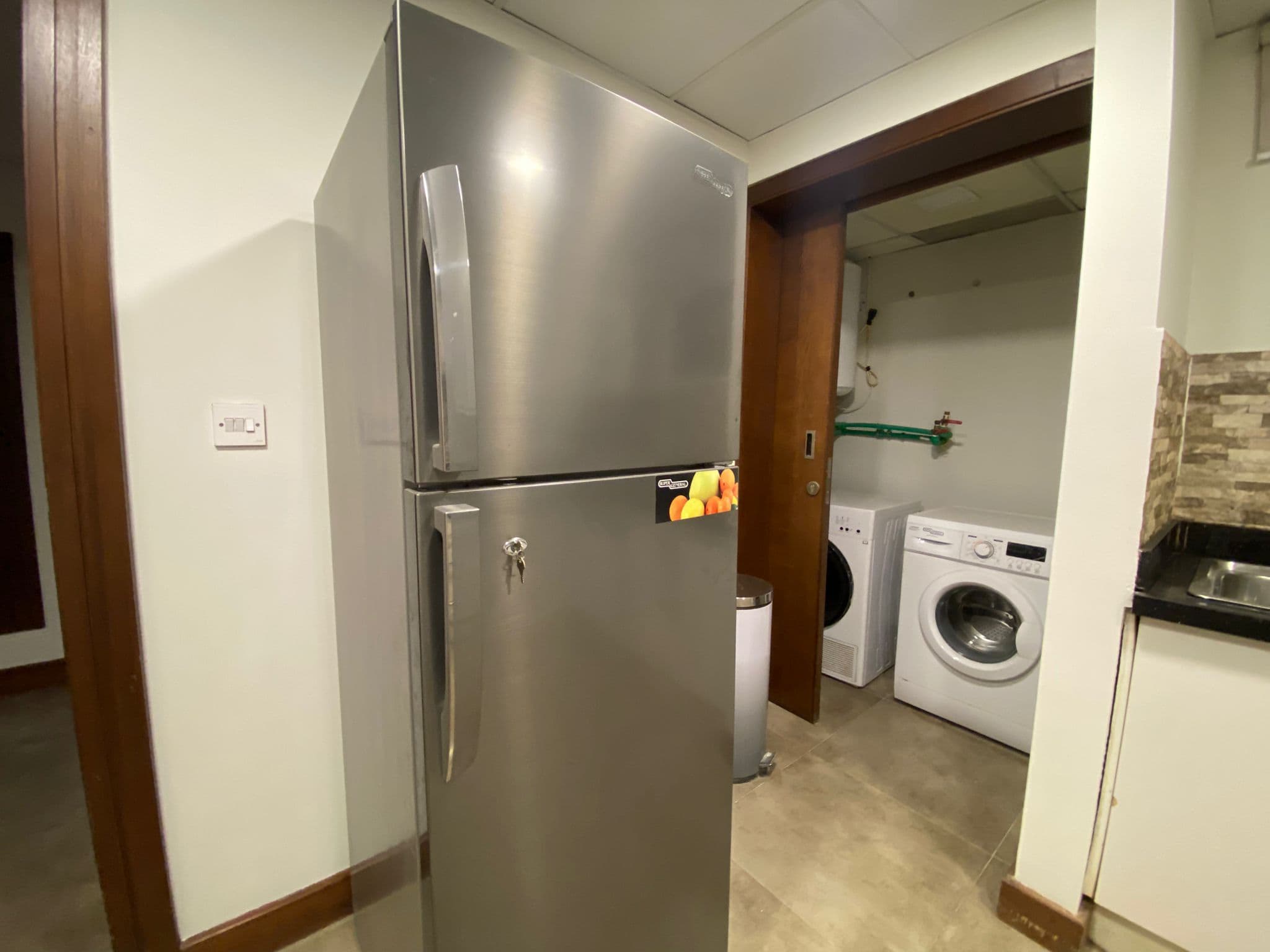 A stainless steel double-door refrigerator is placed next to a laundry area with a washing machine and a countertop in a modern indoor setting.