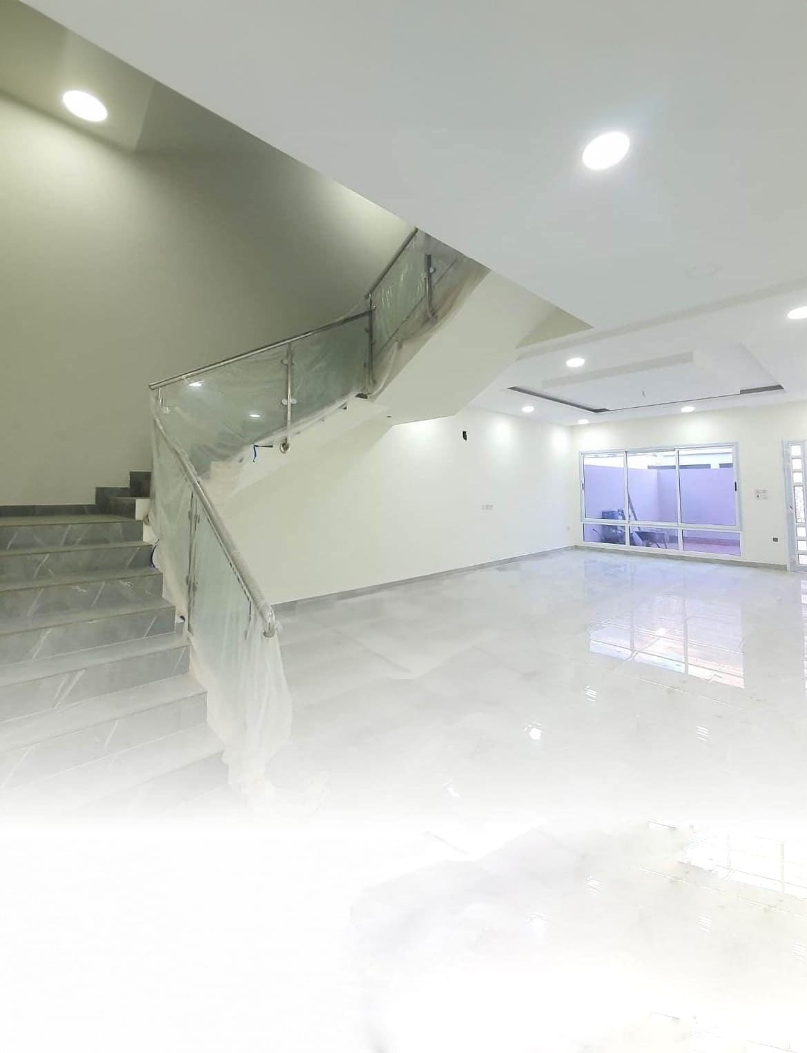 Modern interior with a polished marble floor, a staircase with a glass railing on the left, and a glass-walled room in the background under bright ceiling lights.