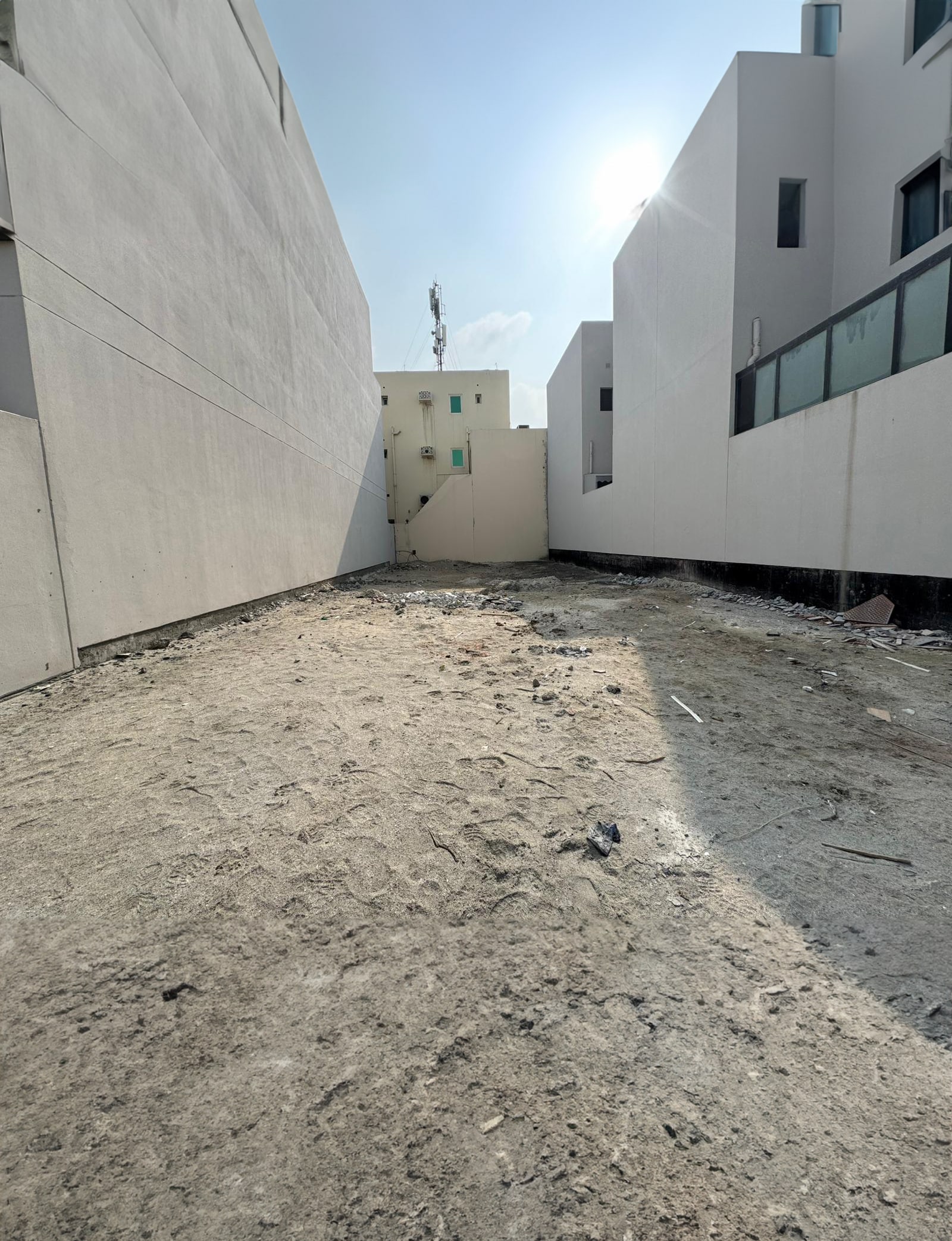 A narrow, empty alleyway with sandy ground between two white buildings under a clear sky.