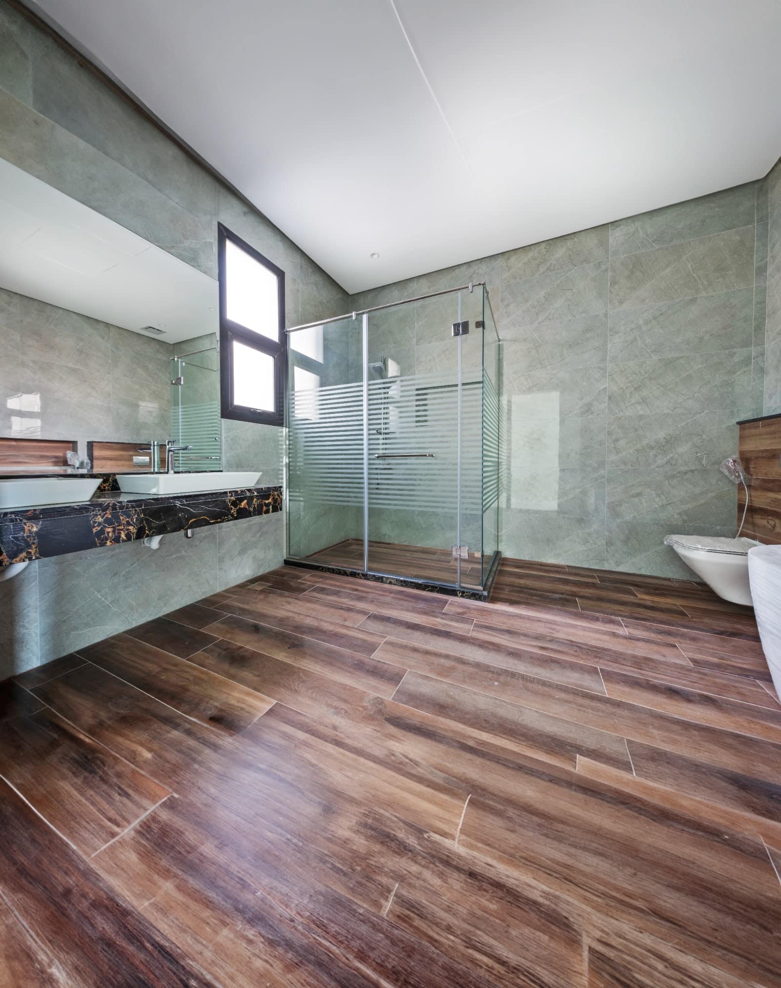 Modern bathroom with large glass shower, wood-look flooring, wall-mounted sink, and toilet.