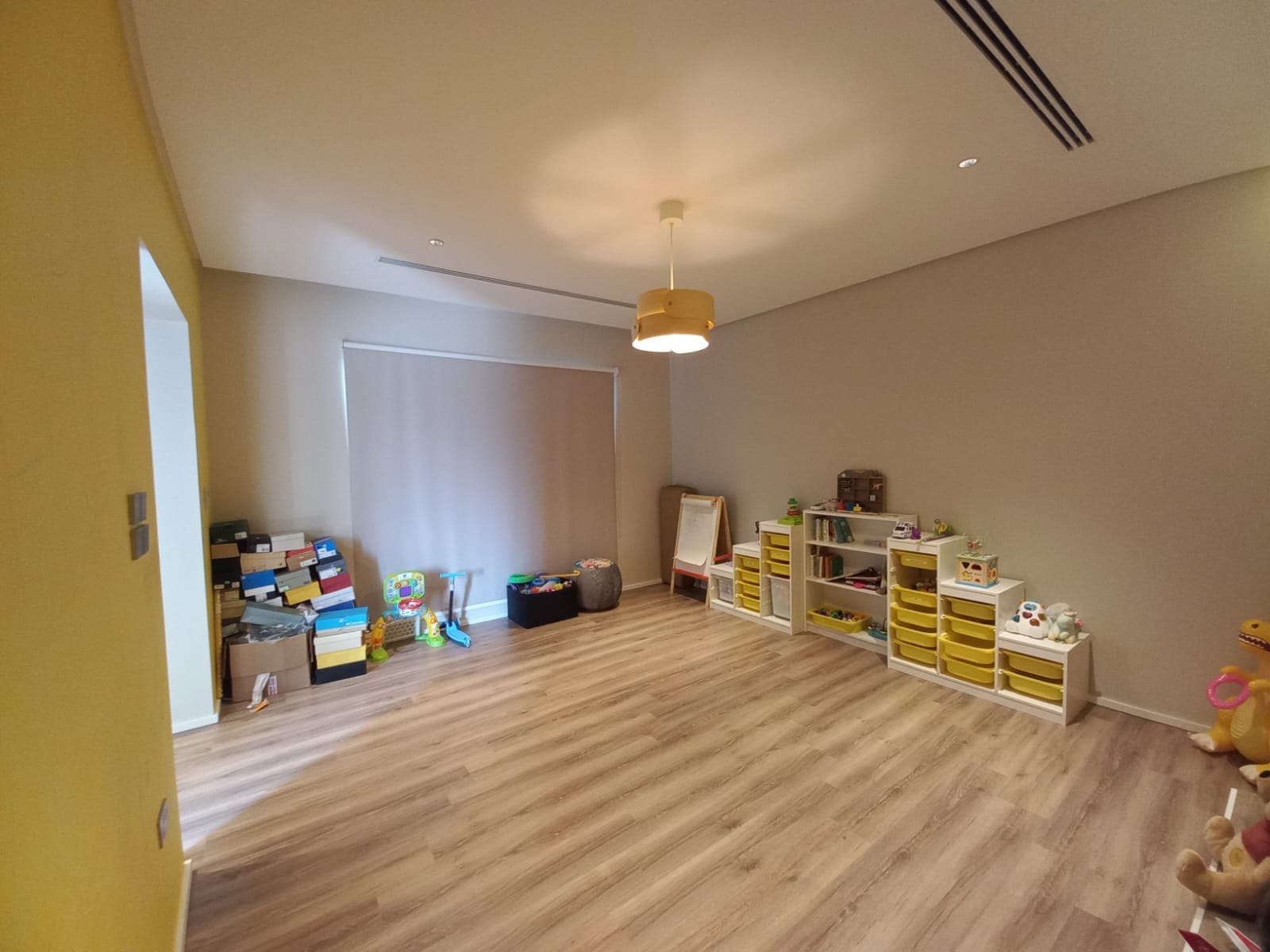 A spacious playroom with wooden flooring, toys, and storage units along the walls. A soft yellow wall and a central ceiling light create a warm atmosphere.