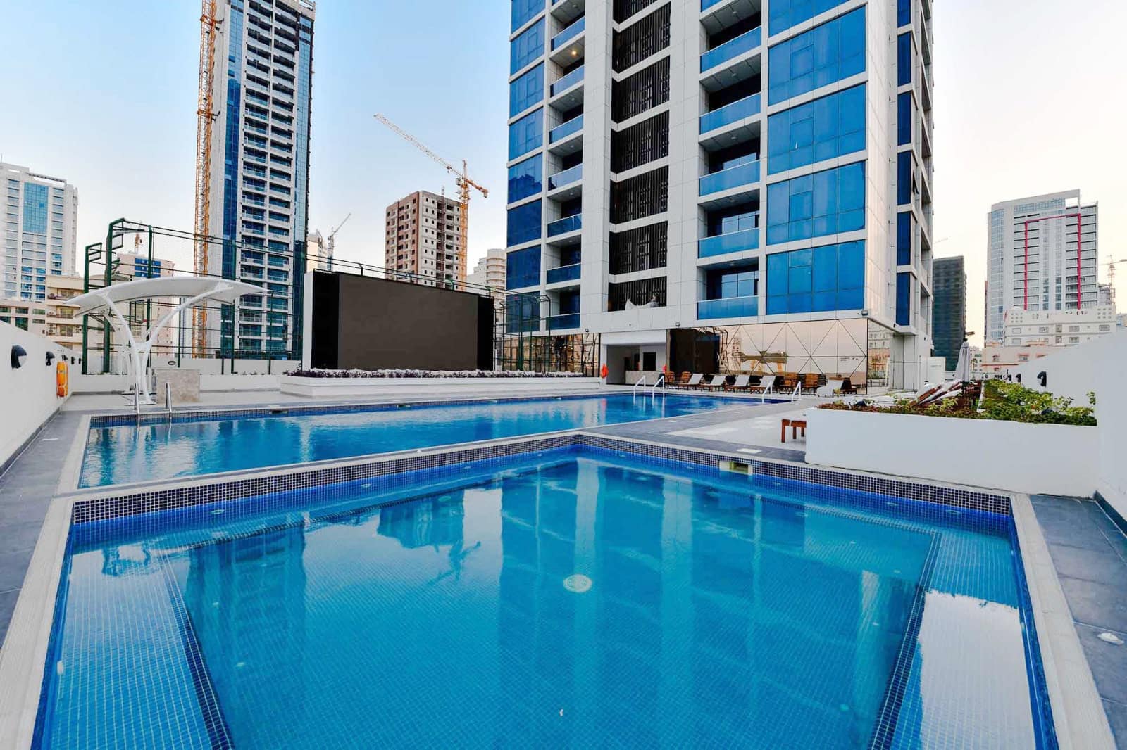 Rooftop swimming pools with clear blue water are surrounded by high-rise buildings and construction cranes. Lounge chairs are arranged on the deck.