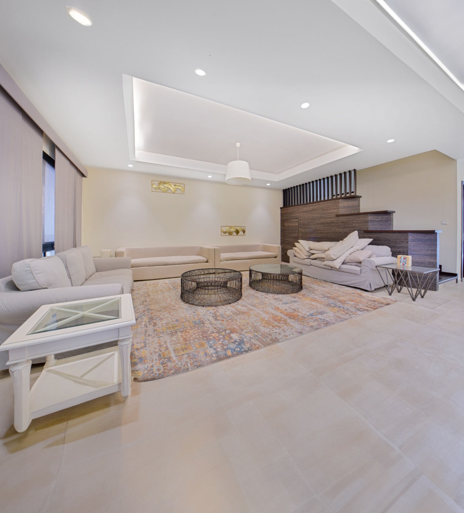 Spacious modern living room with beige sofas, two black wire coffee tables, and a colorful rug. Steps lead up to a mezzanine level. Ceiling lights and a pendant lamp provide illumination.