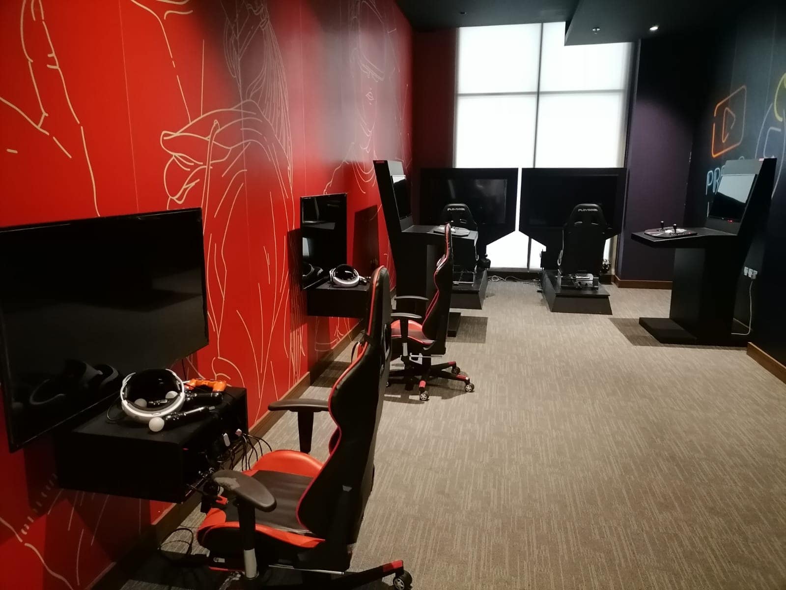 A room with gaming chairs, steering wheels, and monitors, set up for simulation gaming along red and black walls.