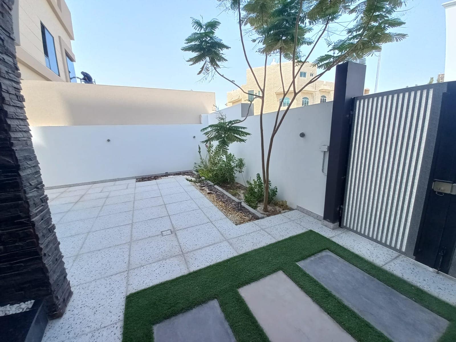 Small outdoor patio with tiled floor, grass patches, a few plants, and a tree. Surrounded by a white wall and metal gate.