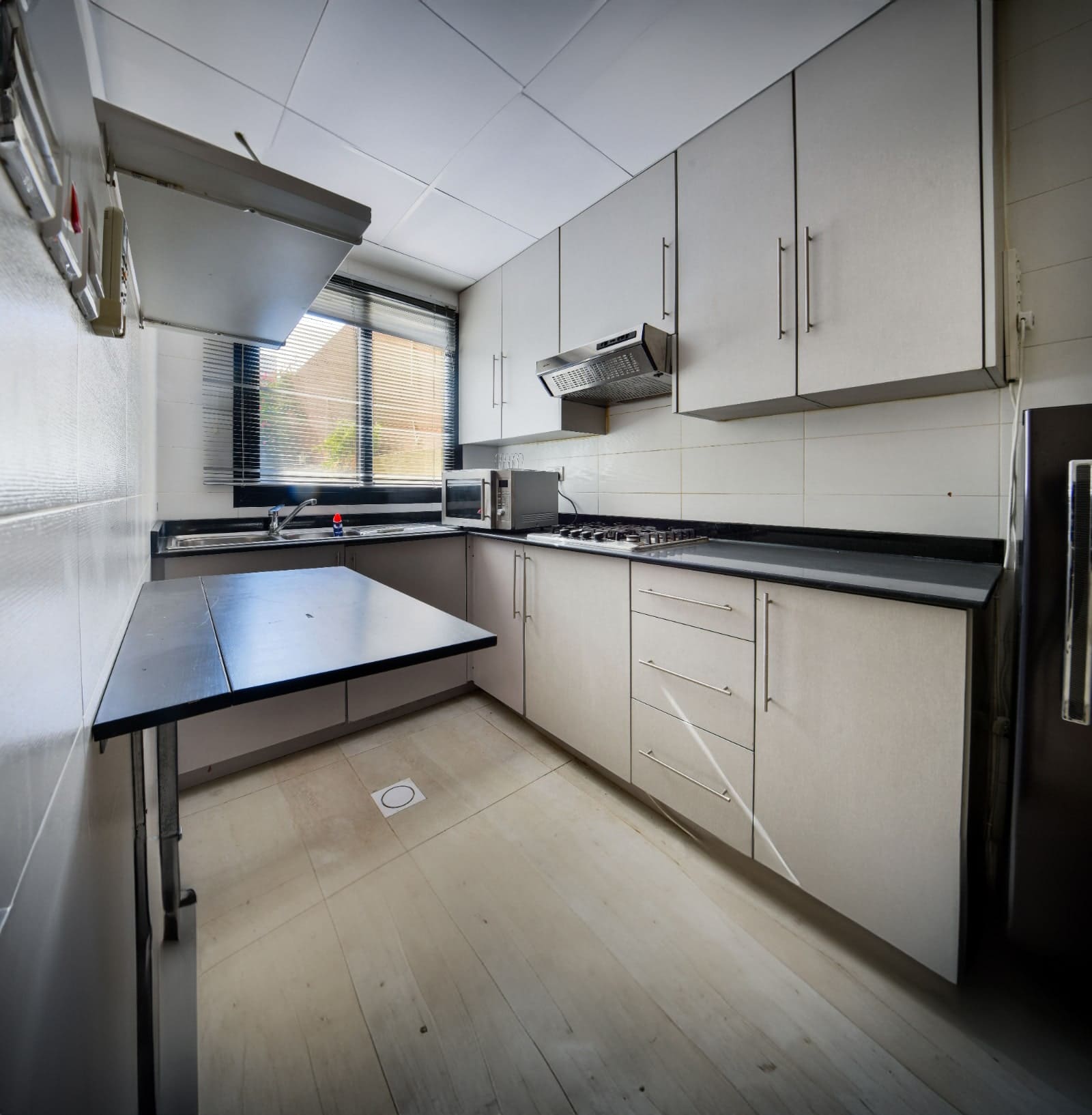 A modern kitchen with gray cabinets, a black countertop, a gas stove, a range hood, and a microwave under a window with blinds. The tiled floor and ceiling have a light finish.