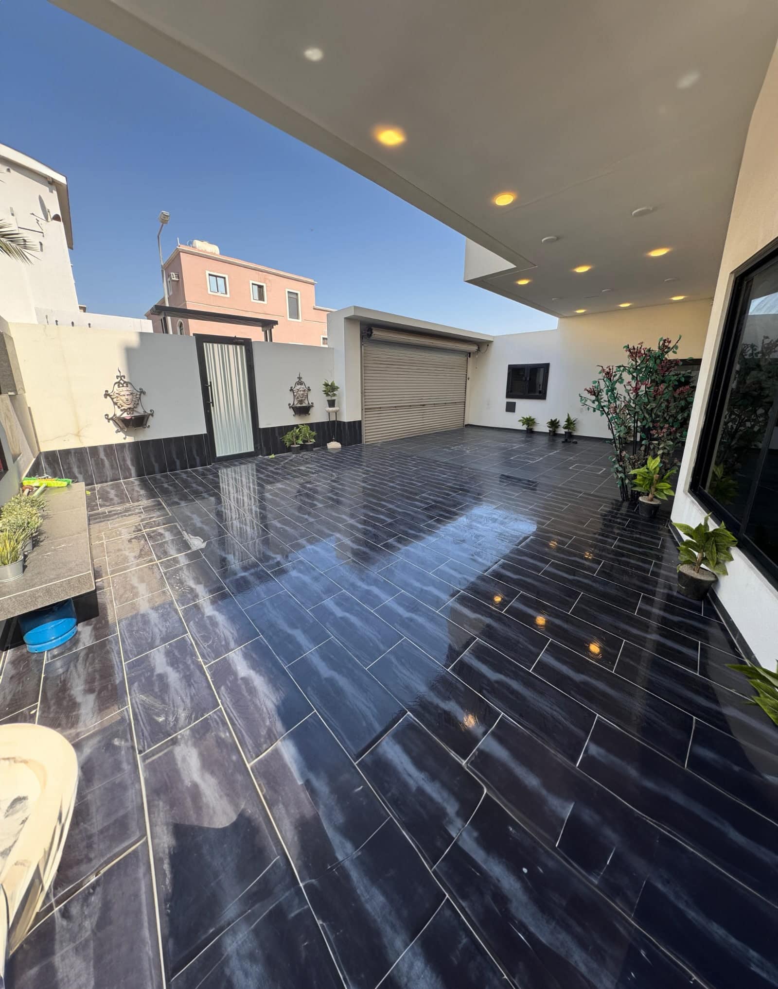 Modern home courtyard with glossy black tiles, potted plants, and partially visible sky. White walls and ceiling with recessed lights, garage door, and gated entrance in the background.