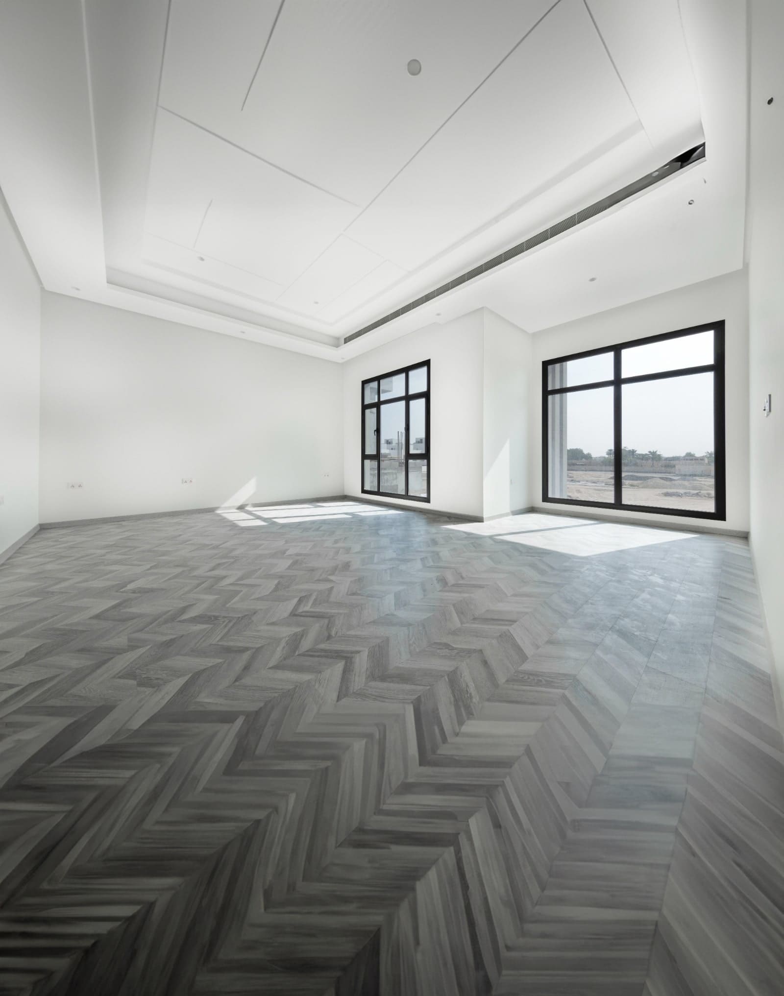 Spacious empty modern room with herringbone-patterned flooring, large windows, and white walls.