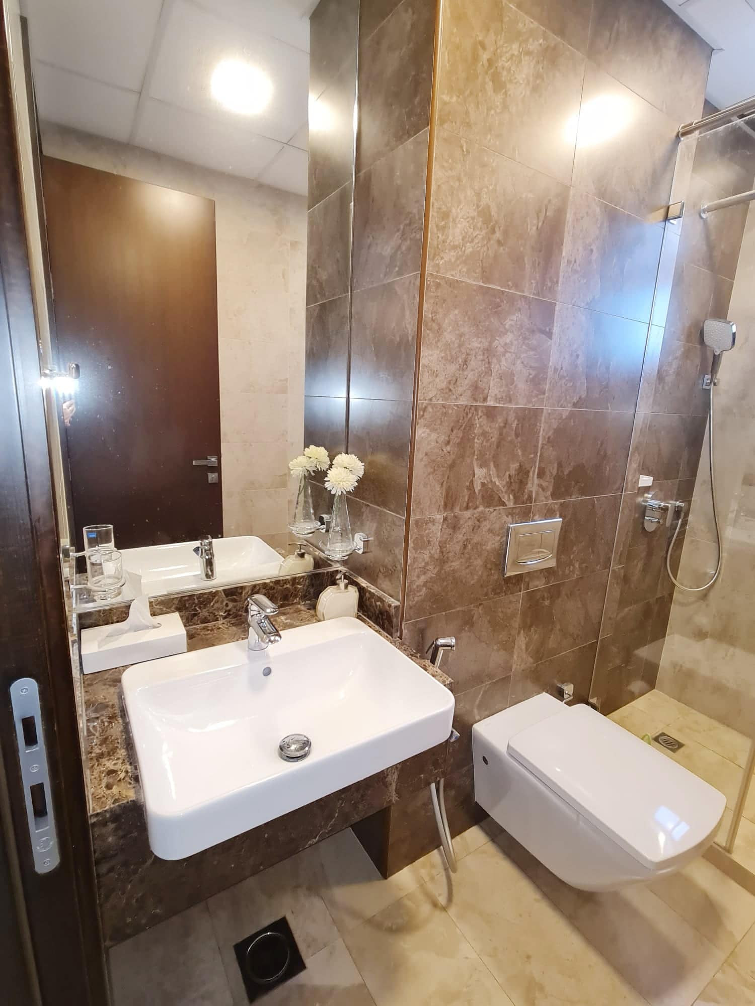 Modern bathroom with a white sink, wall-mounted toilet, and glass shower. Brown marble tiles and a vase with white flowers are on the countertop.