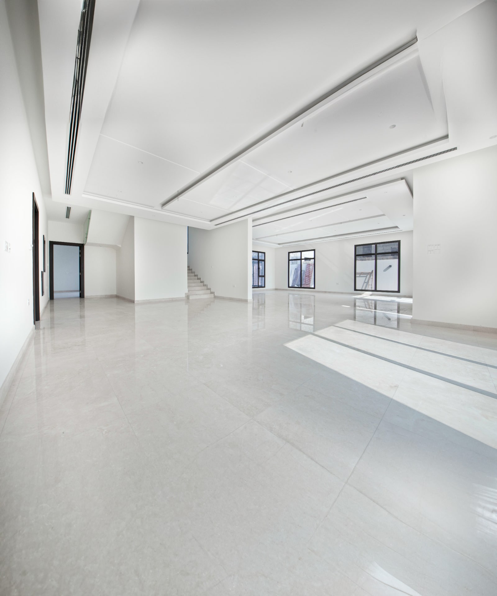 A spacious, empty room with a sleek marble floor, white walls, multiple windows, and a staircase in the corner.