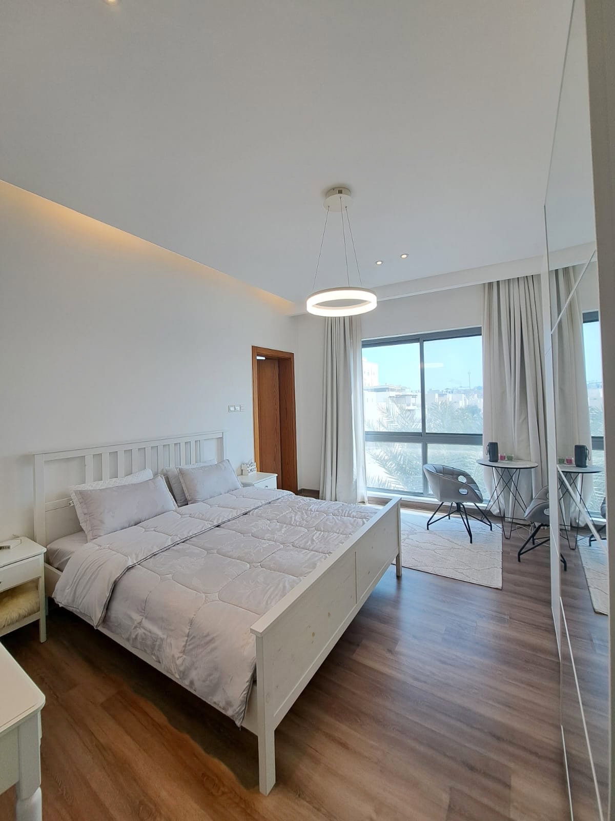 Modern bedroom with a white double bed, large window, wooden floor, and a small desk with chairs. A round ceiling light hangs above, and a sliding closet is on the right.