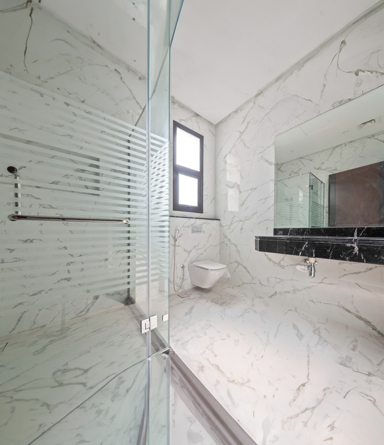 Modern bathroom with marble walls and floor, glass shower enclosure, wall-mounted toilet, and a black countertop with sink.