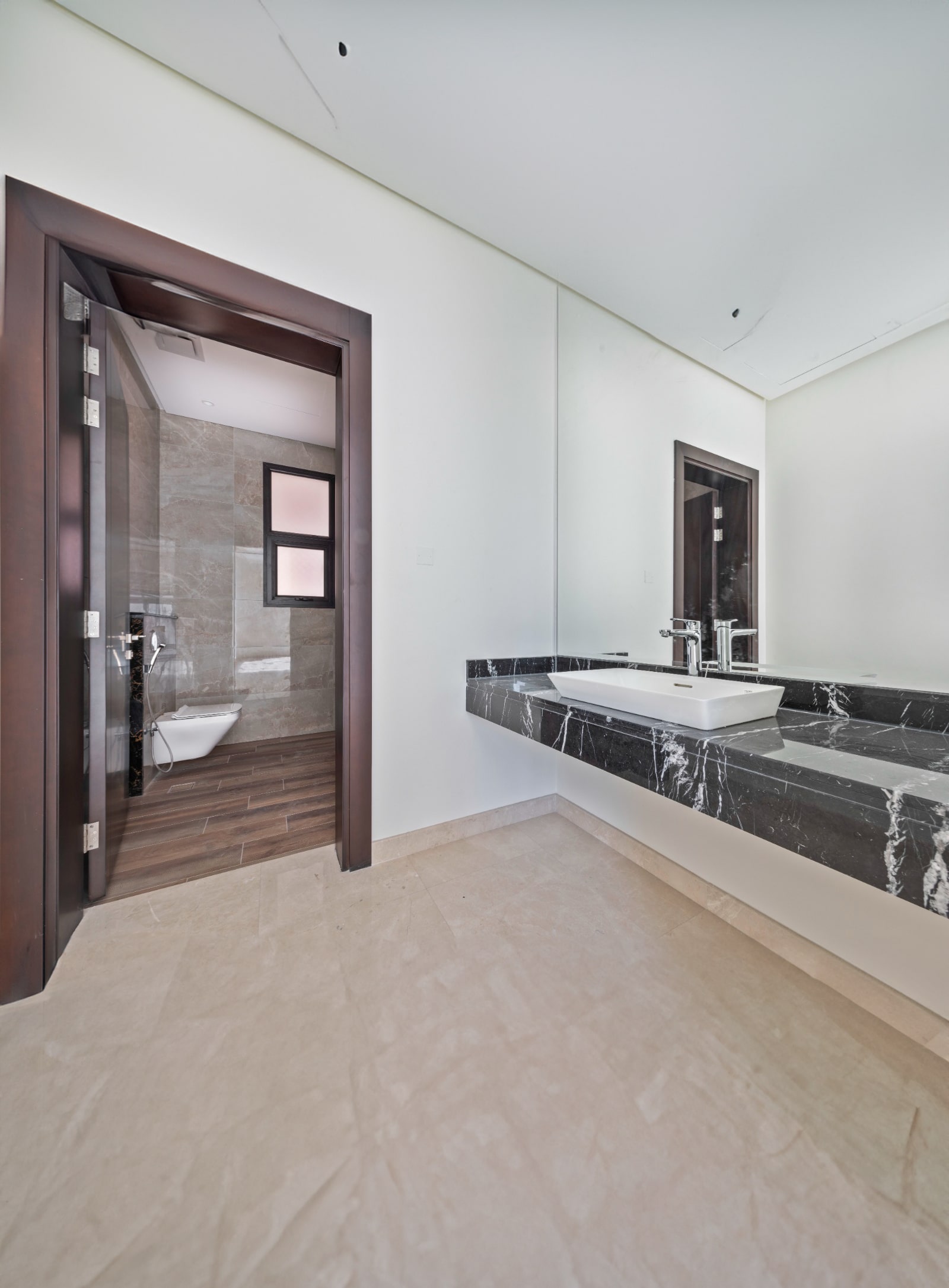 Modern bathroom with beige tile floor, large mirror above a black marble sink, and visible entrance to a separate toilet space with a window on the left.