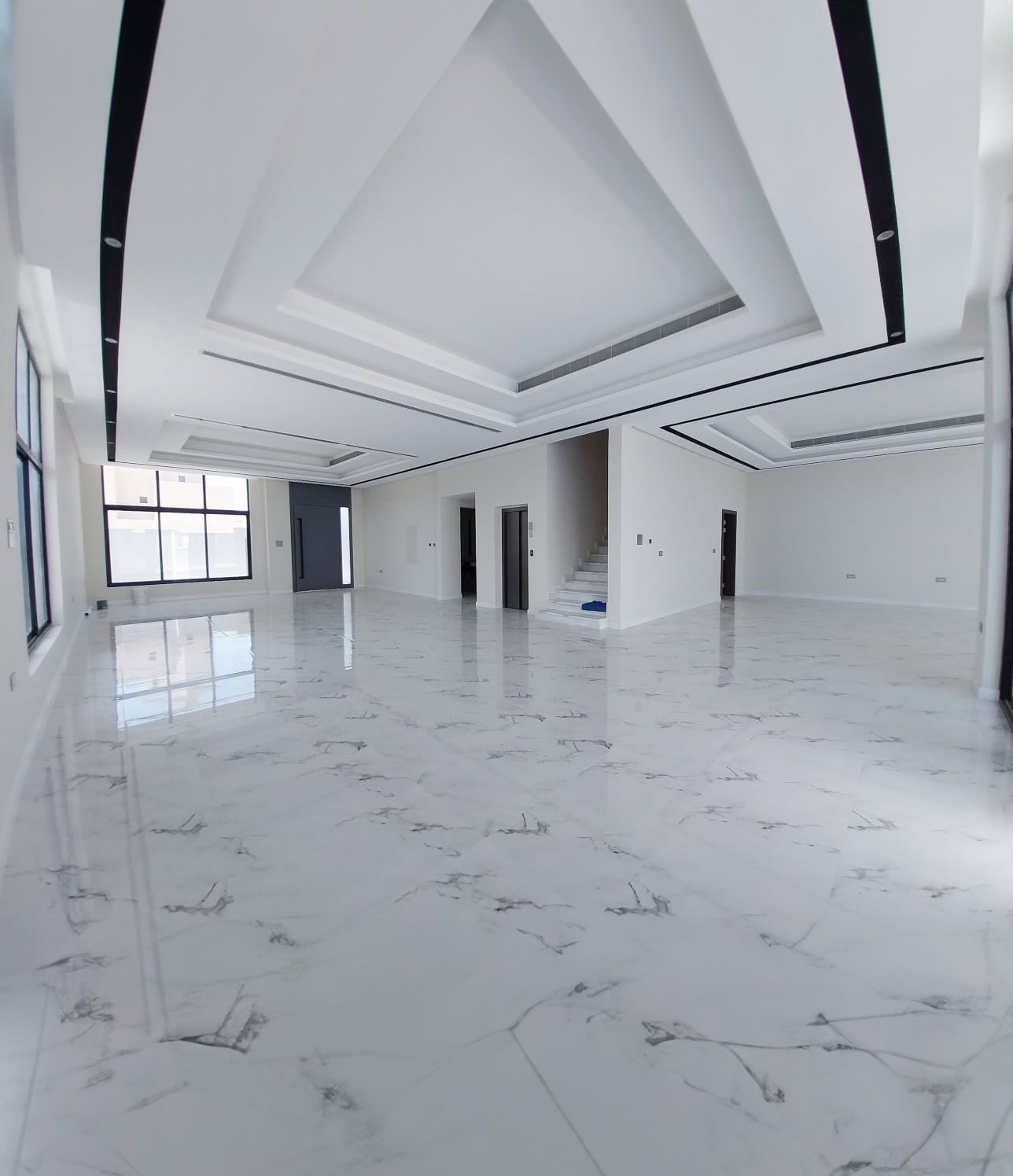 Spacious, empty room with marble-patterned tile flooring, white walls, large windows, and a staircase in the background.