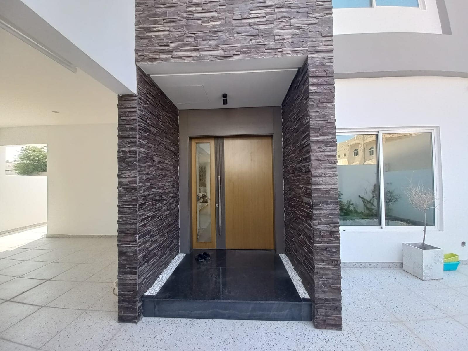 Modern house entrance with a wooden door, flanked by dark stone walls. White exterior with large windows on the right.