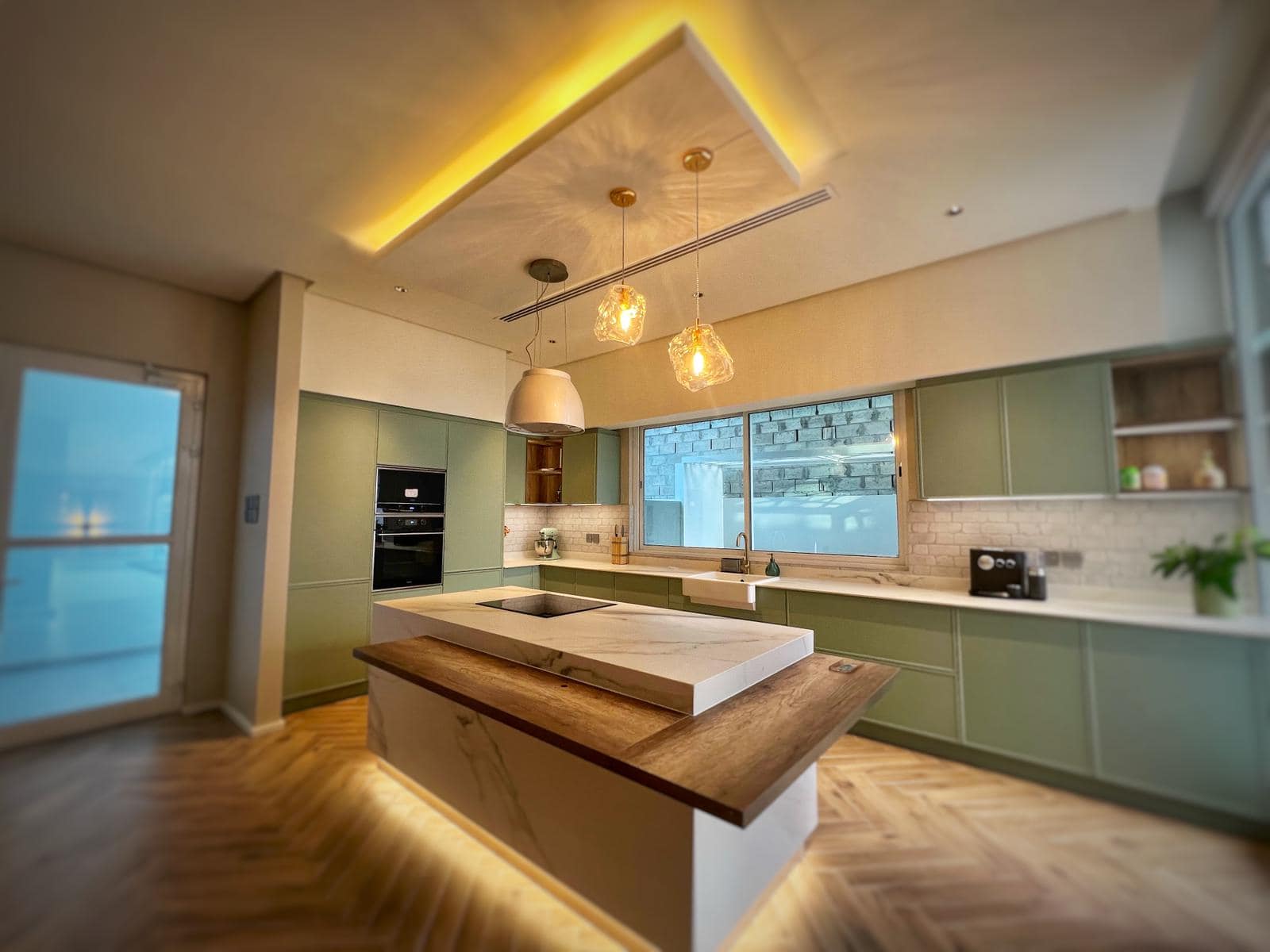 Modern kitchen with a central island featuring a wooden countertop and marble surface. Pendant lights hang above. Cabinets are green, and large windows provide natural light.