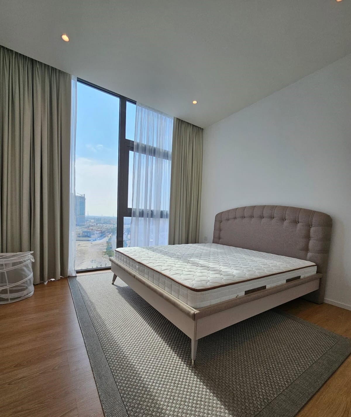 A bedroom with a bed, grey padded headboard, wooden floor, large window with city view, sheer and blackout curtains, laundry basket, and a textured rug.