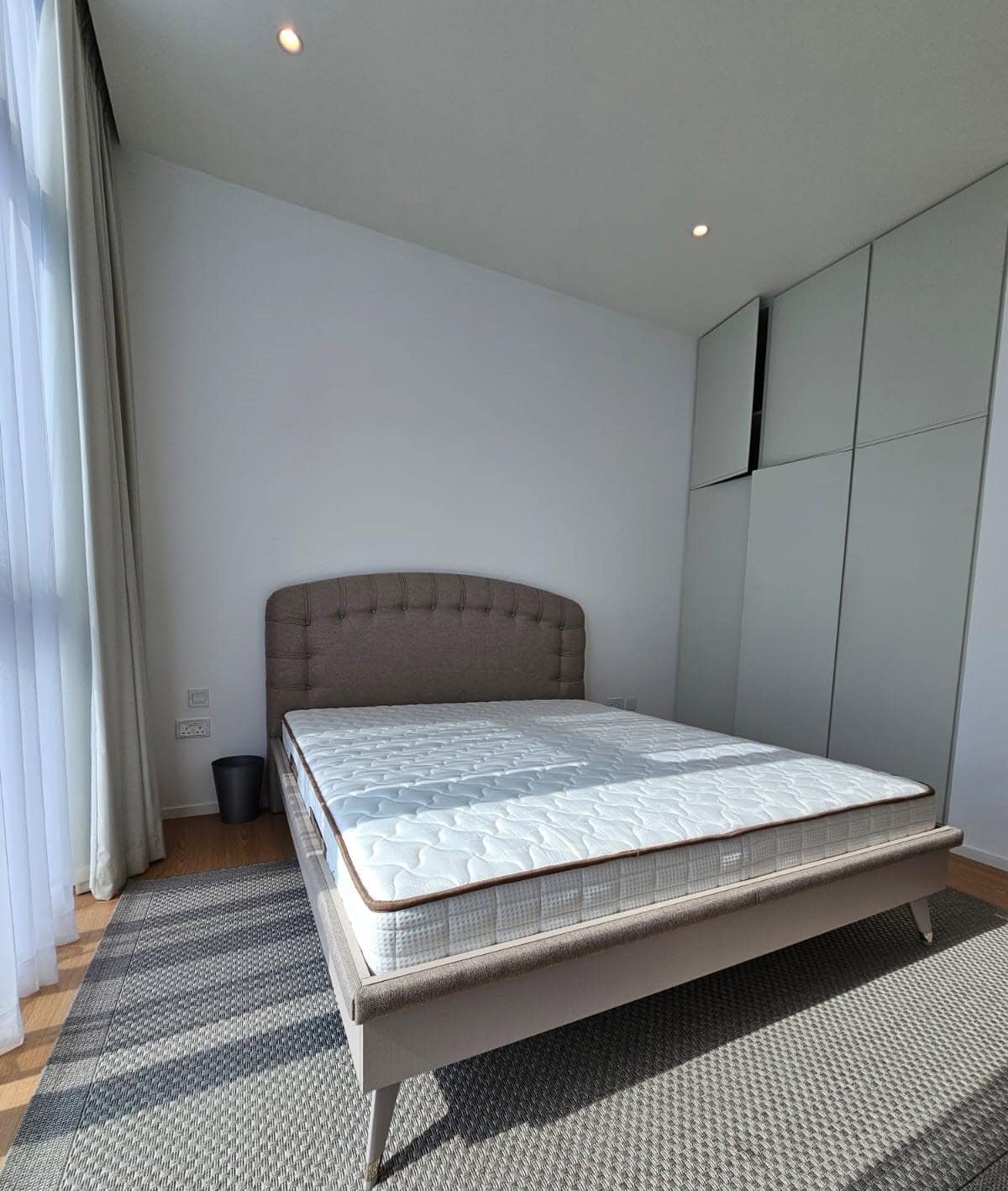 A bedroom with a double bed featuring a tufted headboard, placed on a gray rug. The room has large windows with sheer curtains, a black trash can, and white built-in wardrobes.