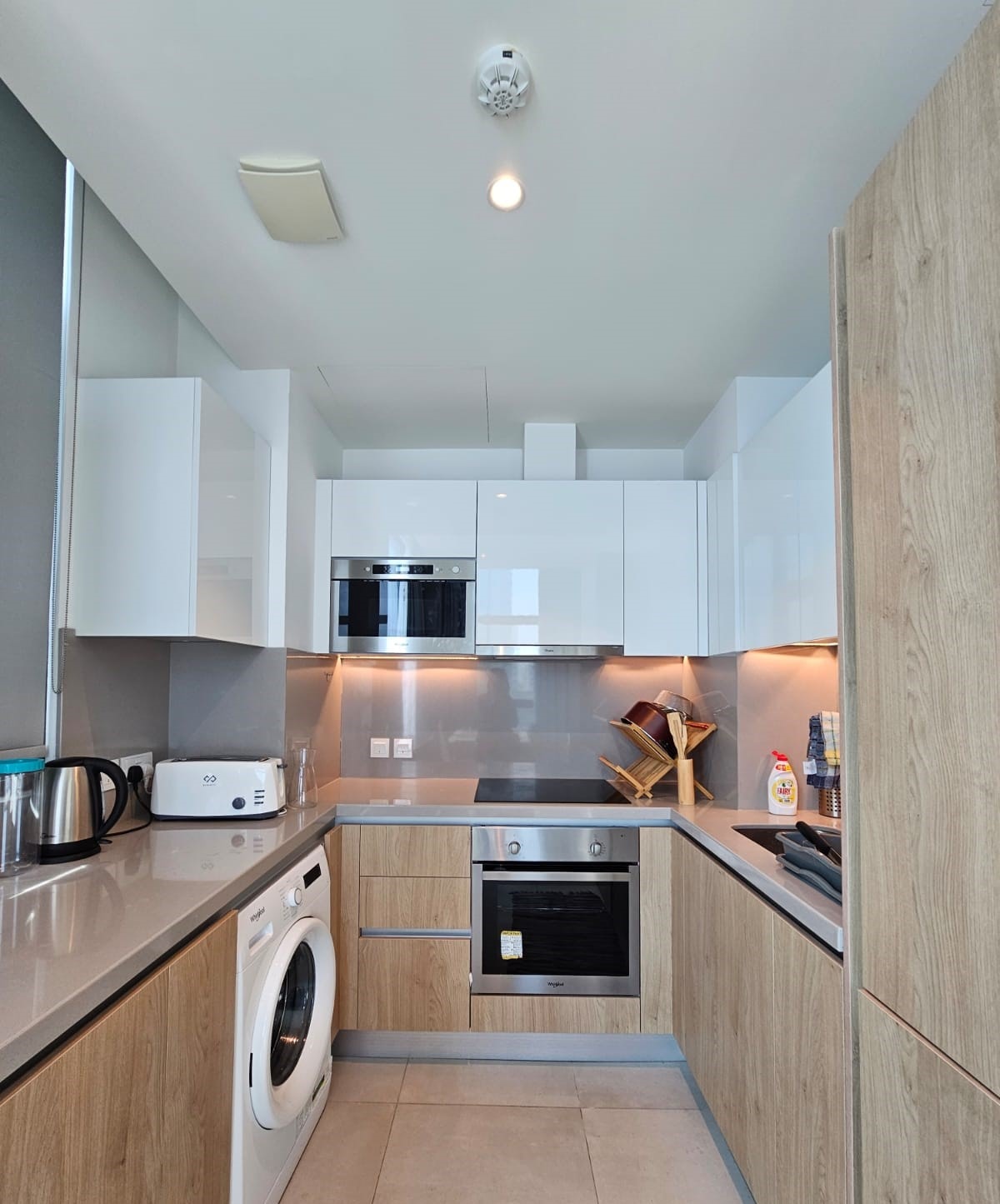 Modern kitchen with light wood cabinets, white appliances, and a sleek countertop. Features built-in oven, microwave, washing machine, toaster, and electric kettle.