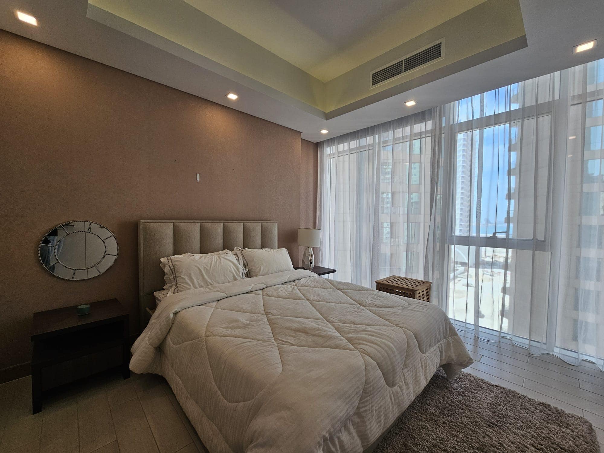 A neatly made bed with white linens in a modern bedroom. Large windows with sheer curtains allow natural light to fill the room. A small round mirror and bedside tables are visible.