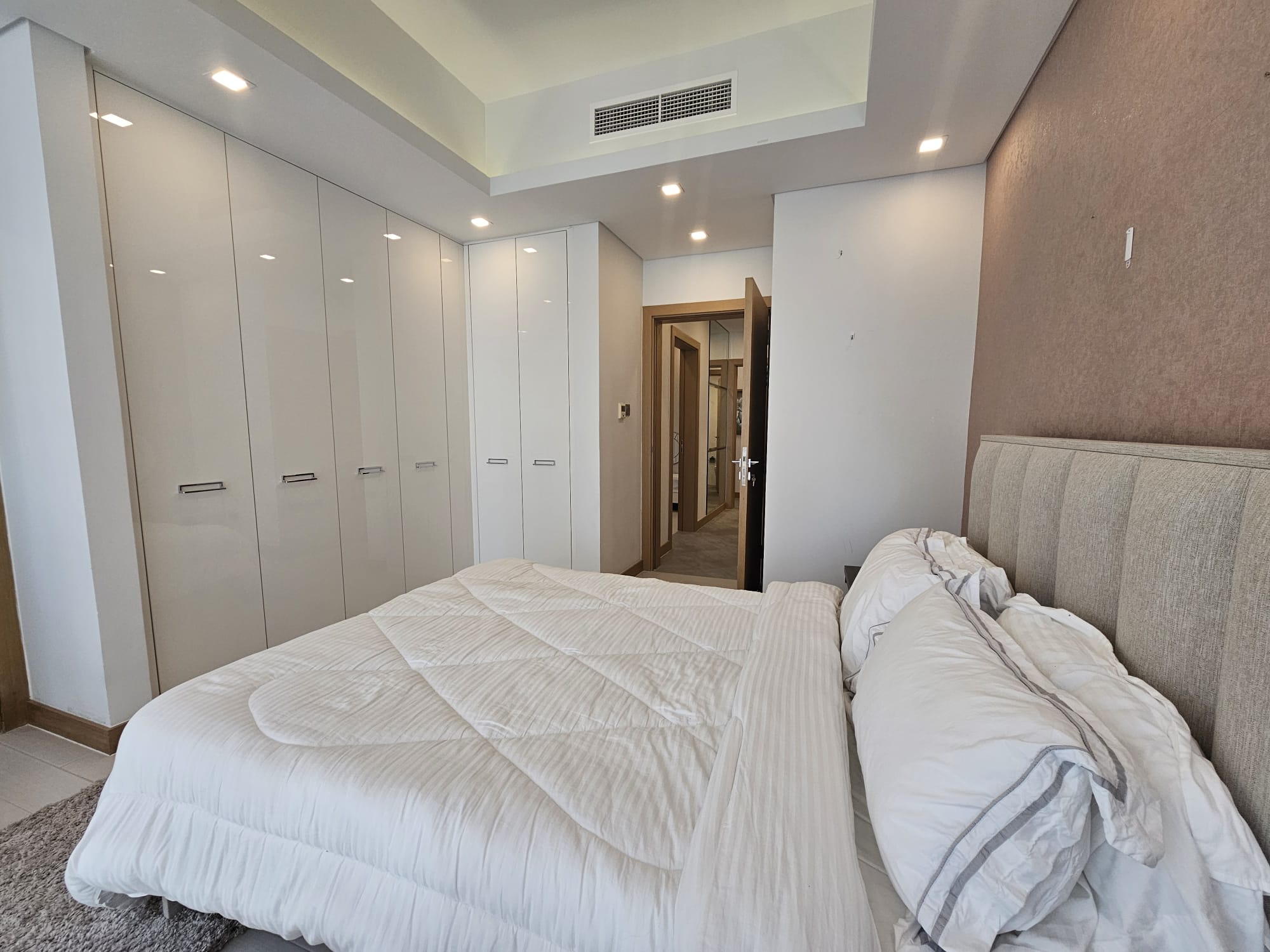 A modern bedroom with a beige upholstered bed, white bedding, built-in wardrobes, recessed lighting, and a carpeted floor. A door leads to an adjacent room.