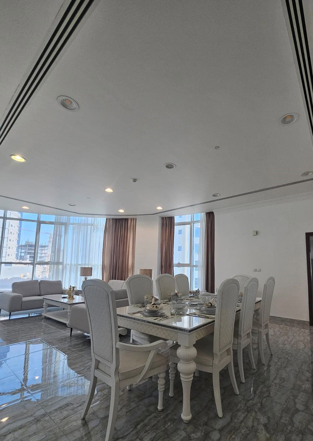 Elegant dining room with a long table set for eight, white chairs, and a corner seating area. Large windows with sheer curtains and city view. Marble floor and modern lighting fixtures.