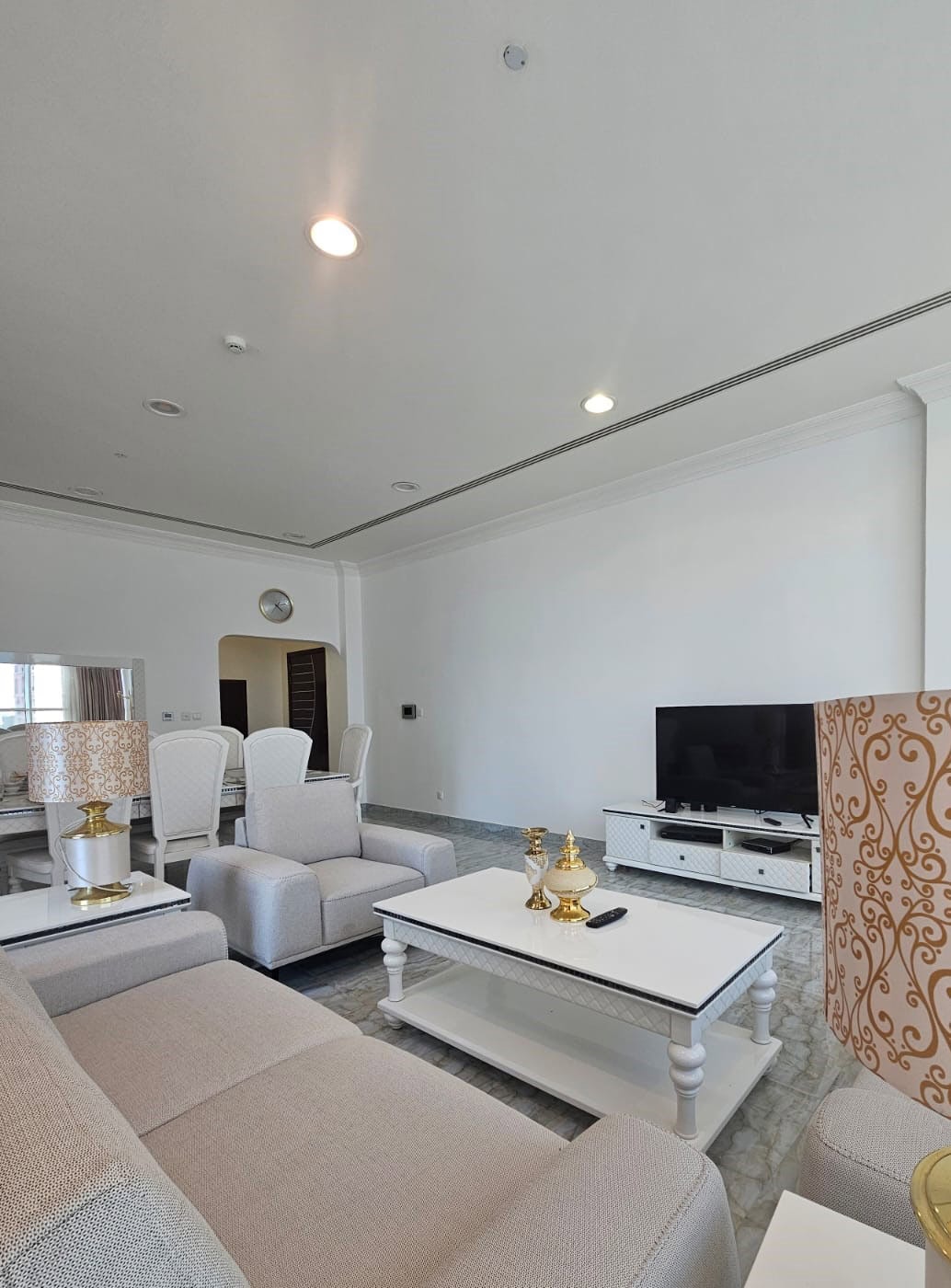 Spacious living room with beige sofas, a white coffee table, gold decor, a large flat-screen TV on a white stand, and dining table with high-back chairs in the background.