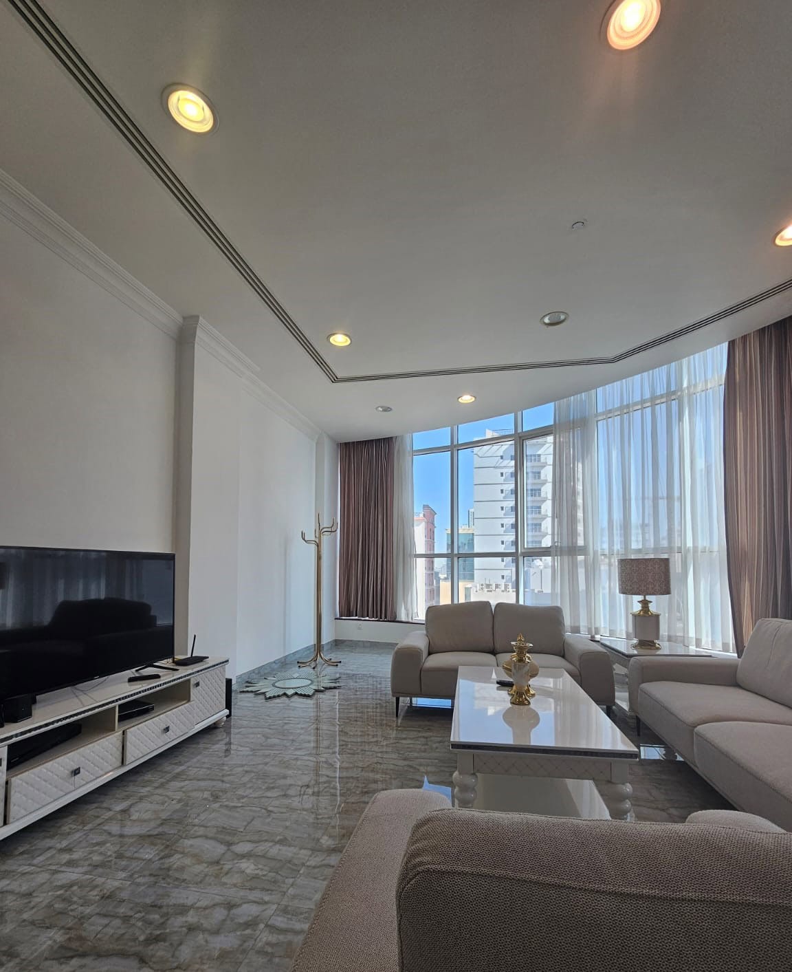 Modern living room with large windows, gray sofas, a glass coffee table, and a TV on a white stand. A coat rack stands near the window, and the floor is marble.