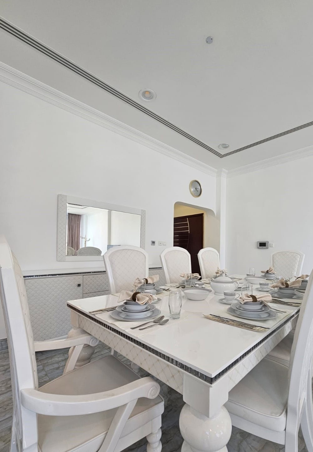 A white dining room with a rectangular table set for six. Plates, bowls, and glasses are neatly arranged. White cushioned chairs surround the table, and a wall clock is visible.