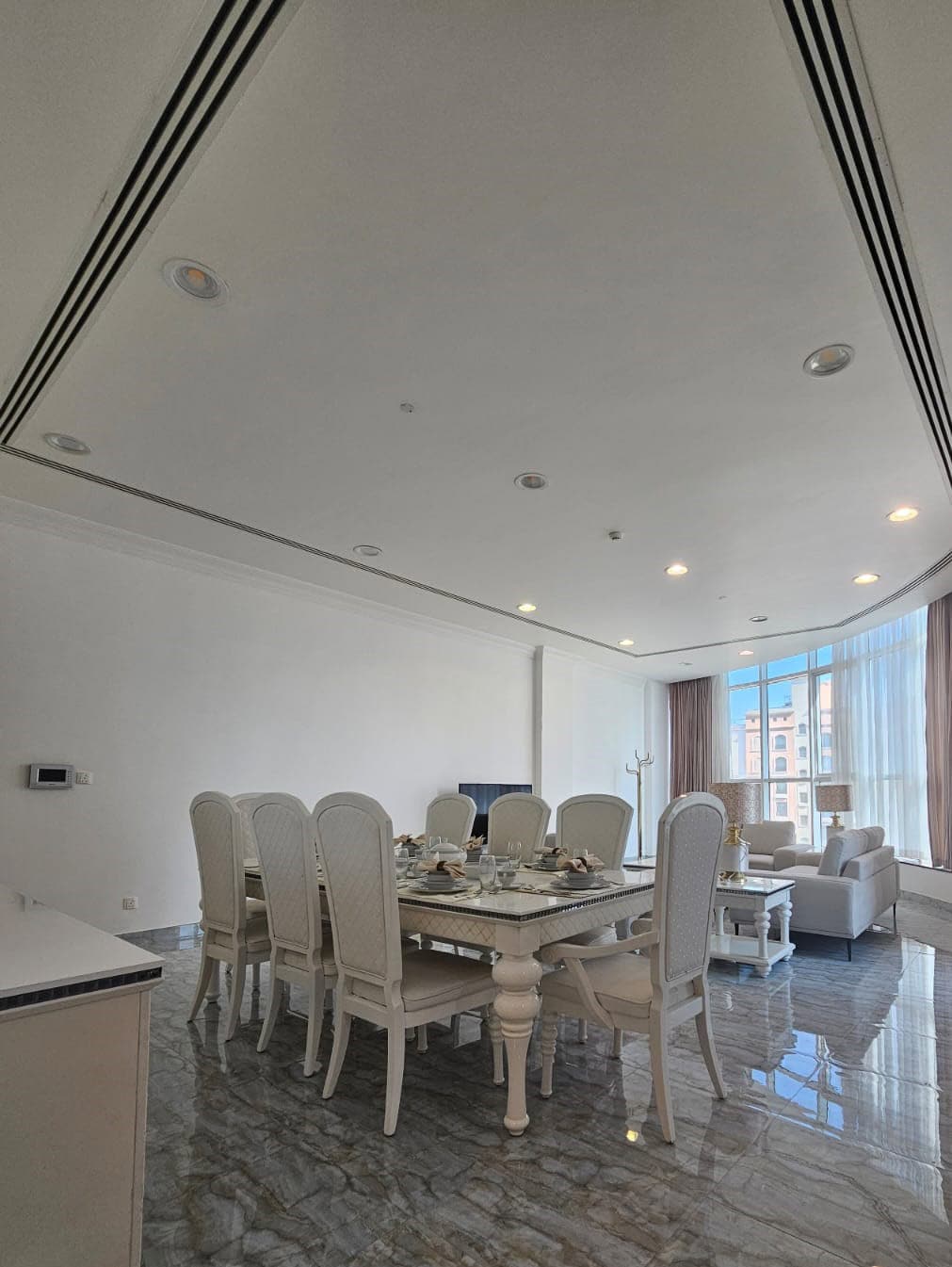 A dining room with a long table and eight chairs, a living area with sofas, large windows, and marble flooring.