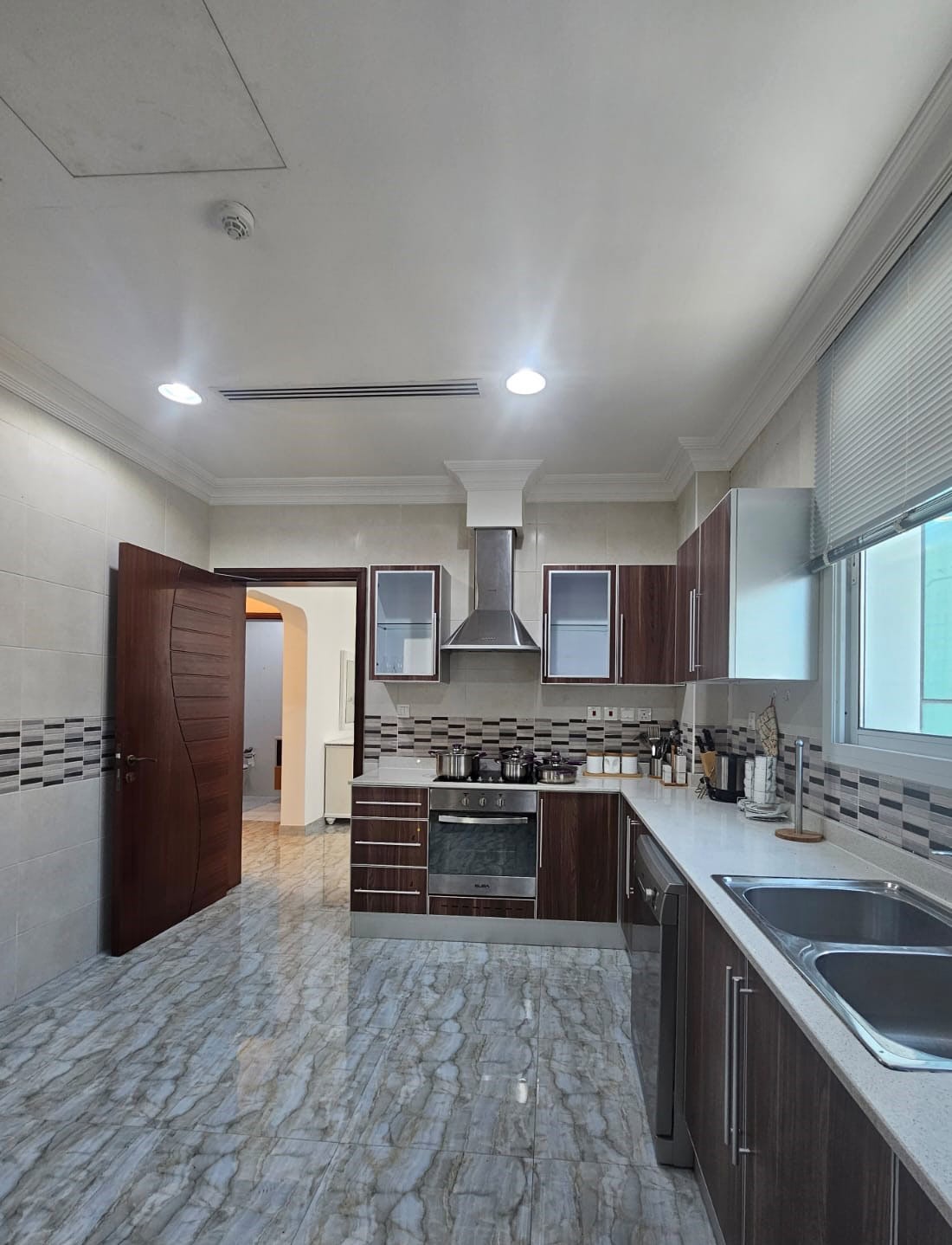 A modern kitchen with brown cabinets, stainless steel appliances, and a tiled floor. Sunlight streams through a window, illuminating the countertop and cookware. An open door leads to another room.