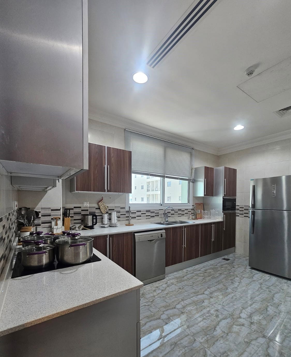 Modern kitchen with stainless steel appliances, wooden cabinets, and marble floor. Pots are on the stovetop, with a dishwasher and fridge nearby. A window provides natural light.