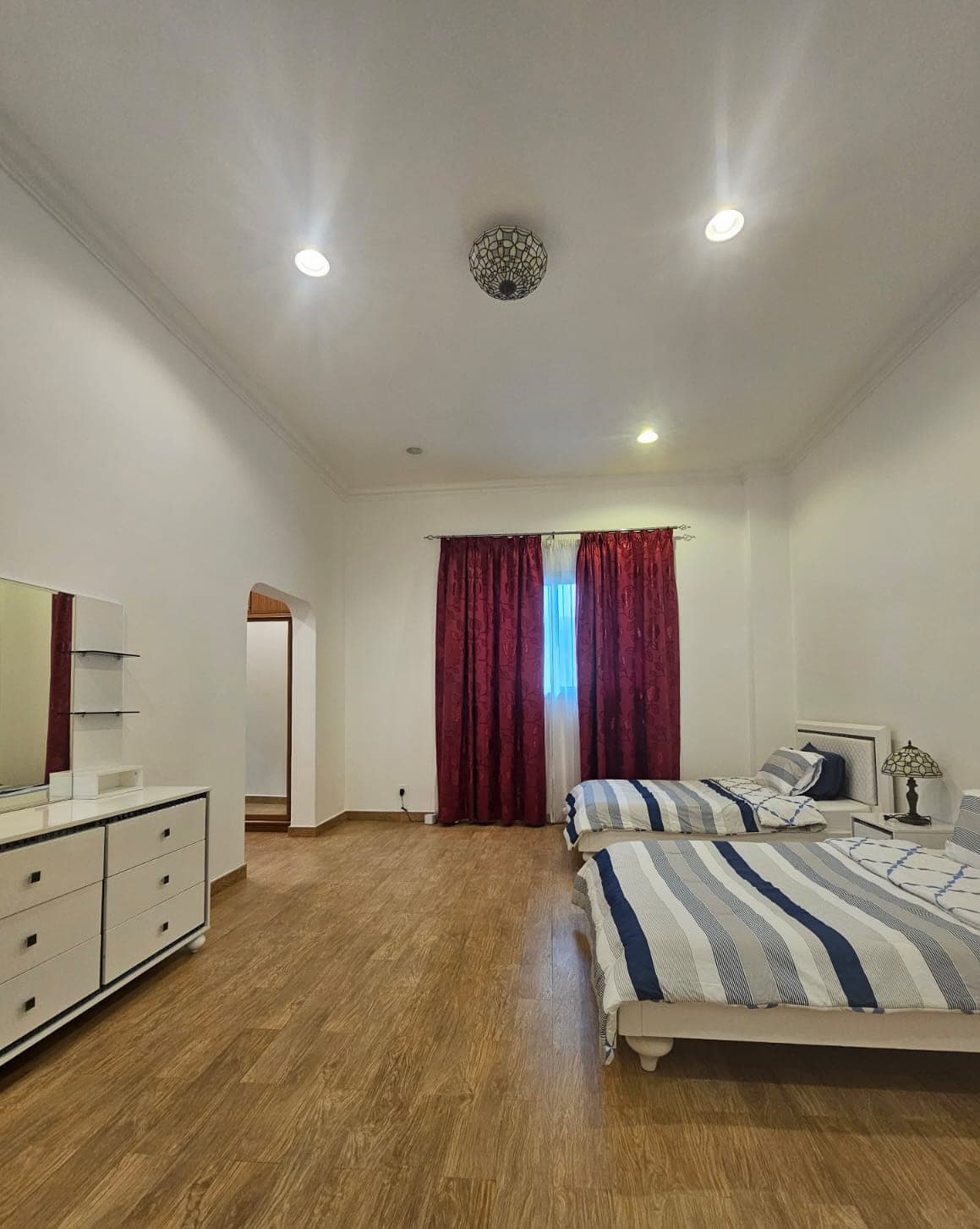 A bedroom with two beds, striped bedding, a white dresser with a mirror, and red curtains covering a window. The floor is wood and the ceiling has recessed lighting.