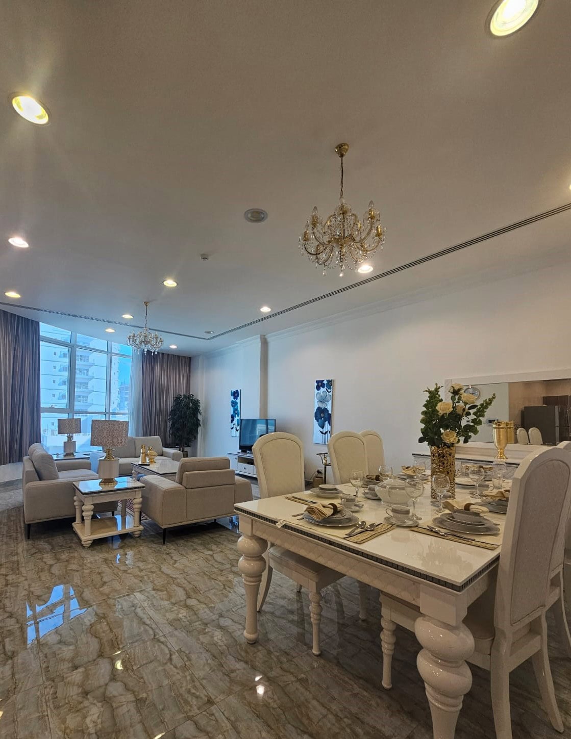 Elegant open-plan living and dining area with beige furniture, marble flooring, and chandeliers. A large window offers natural light, and the table is set for dining.