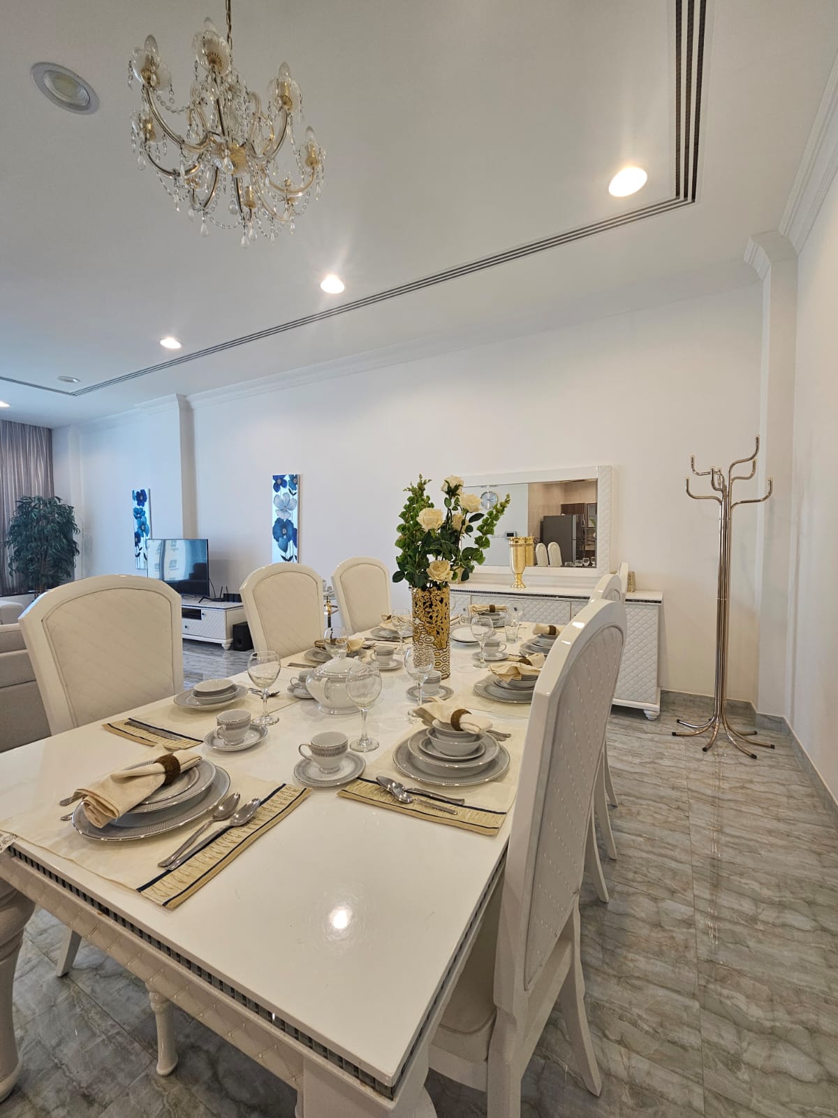 Elegant dining room with a white table set with plates, cups, and cutlery. A chandelier hangs above. A living area is visible in the background with a TV and couches.