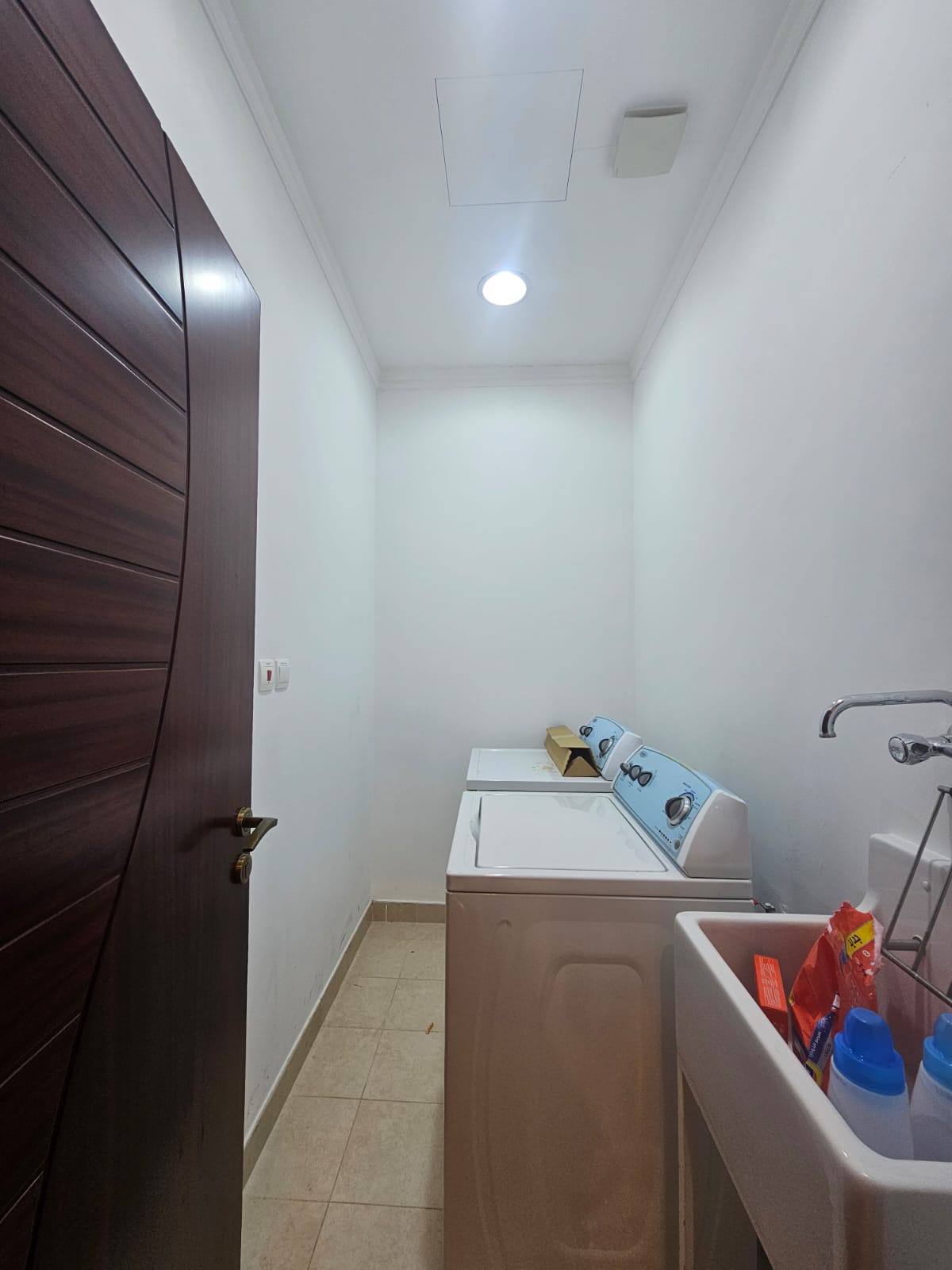 Laundry room with a wooden door, washing machine, dryer, and a sink with detergent bottles on the side. White walls and tiled floor.