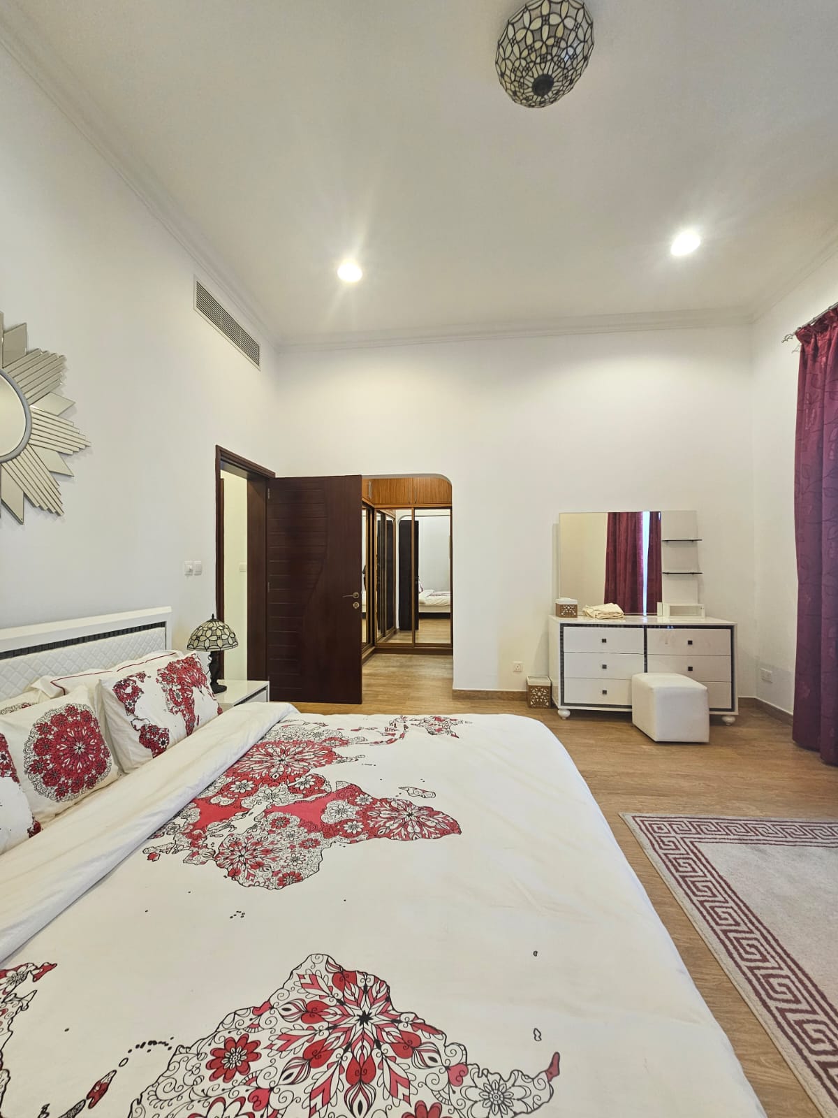 Bedroom with a bed featuring floral bedding, a mirror on a white dresser, a small upholstered stool, and maroon curtains. An open door reveals another room.