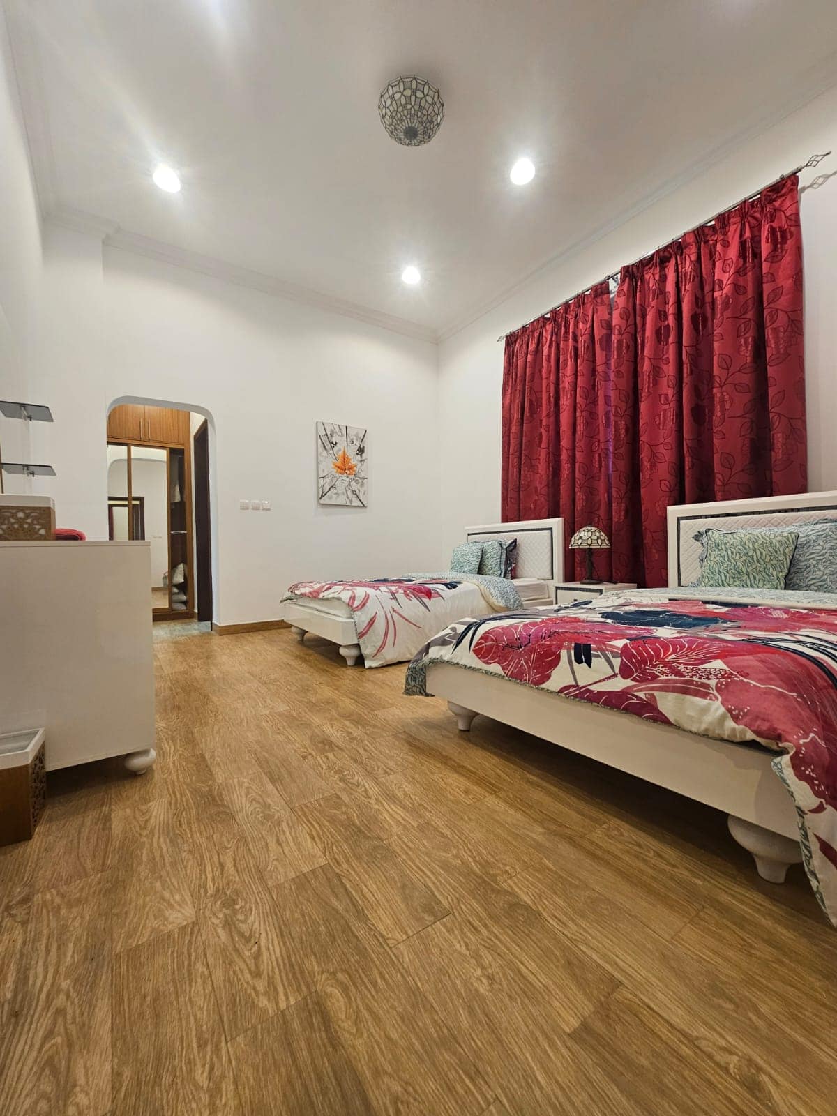 A bedroom with two single beds, colorful bedding, wooden floor, red curtains, a ceiling light, and a wall painting.